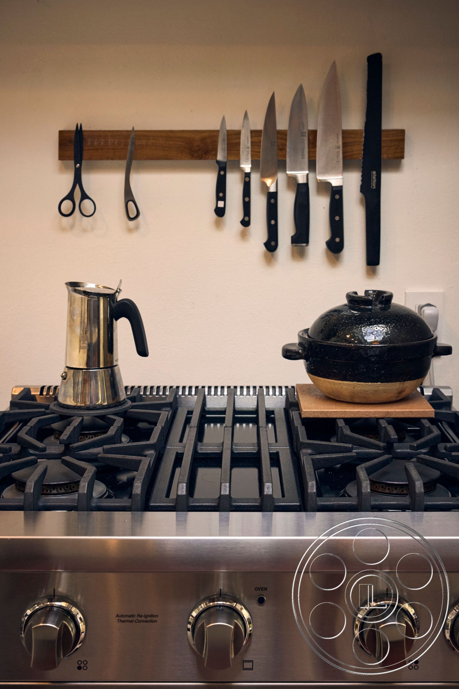 MCM 26 - neutral color palette, open kitchen layout, stainless steel gas range, modern kitchen interior, wooden knife rack, black ceramic pot on wooden board, hanging kitchen knives, metal stovetop grates, industrial kitchen design, minimalist cooking space