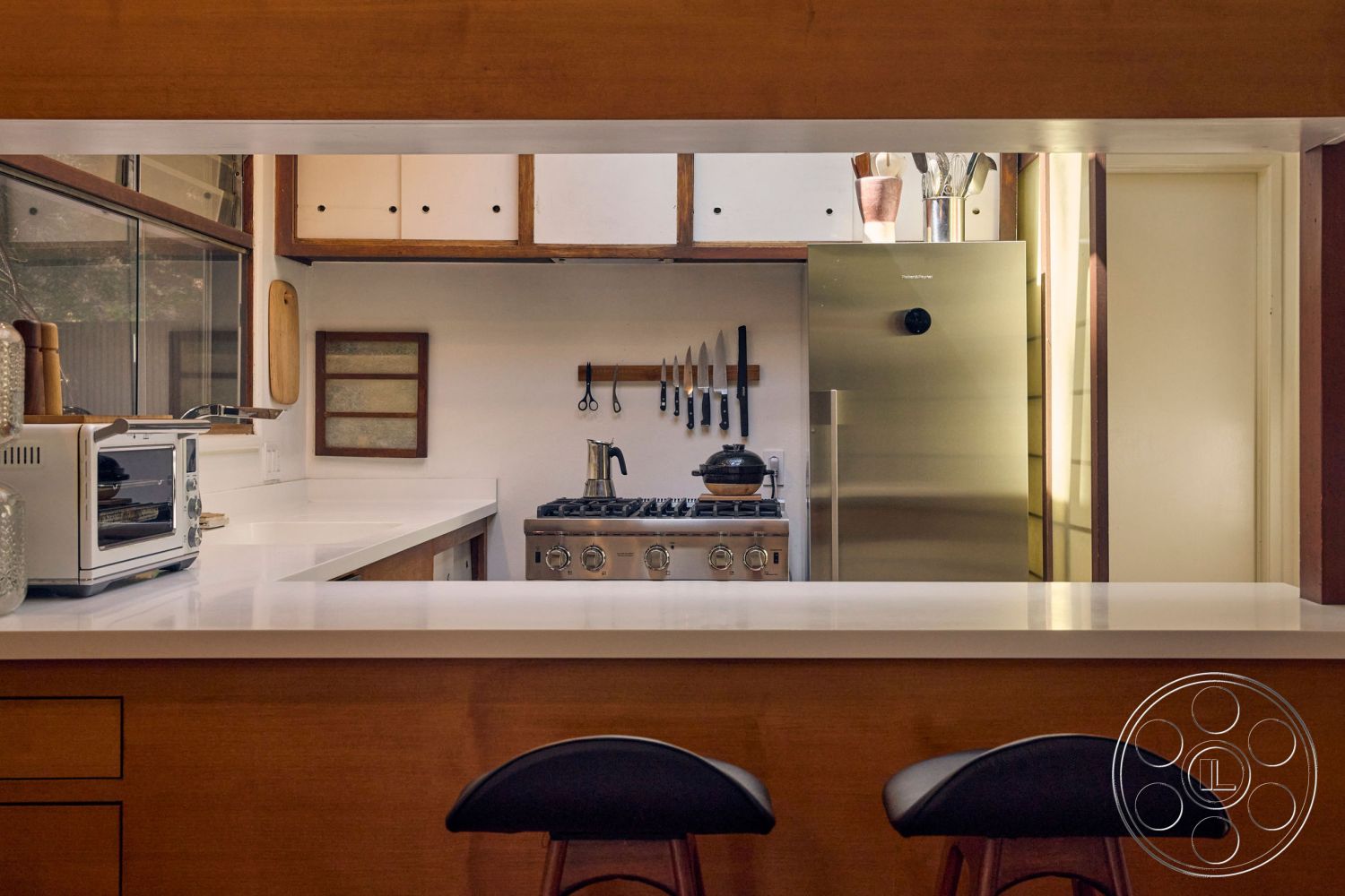 MCM 26 - neutral color palette, stainless steel appliances, open concept layout, modern bar stools, double-hung windows, interior kitchen design, minimalist kitchen decor, white quartz countertops, wood panel cabinetry, kitchen island with seating, contemporary interior lighting, shaker-style cabinet doors, wooden trim accents, knife rack wall hanging, textured kitchen backsplash