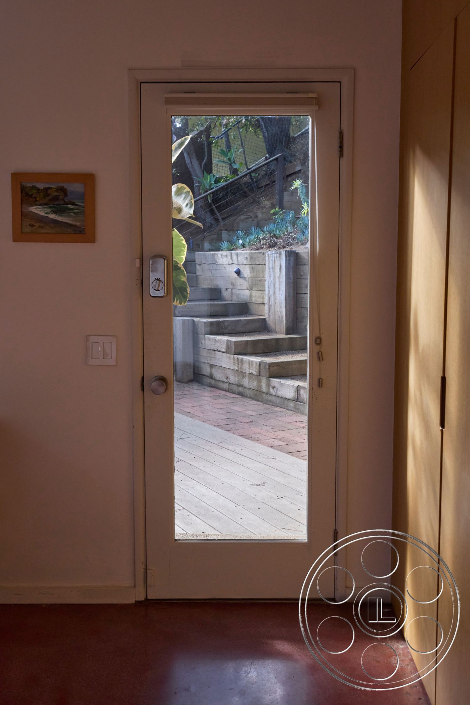 MCM 26 - exterior concrete steps, modern minimalist design elements, interior wooden framed glass door, exposed wood paneling interior walls, red stained concrete flooring, wooden outdoor deck with planters, landscaped outdoor garden, greenery surrounding outdoor stairs, natural light through glass door, sloping hillside property, interior step down transition, wood railing along outdoor steps, colorful wall art in interior foyer