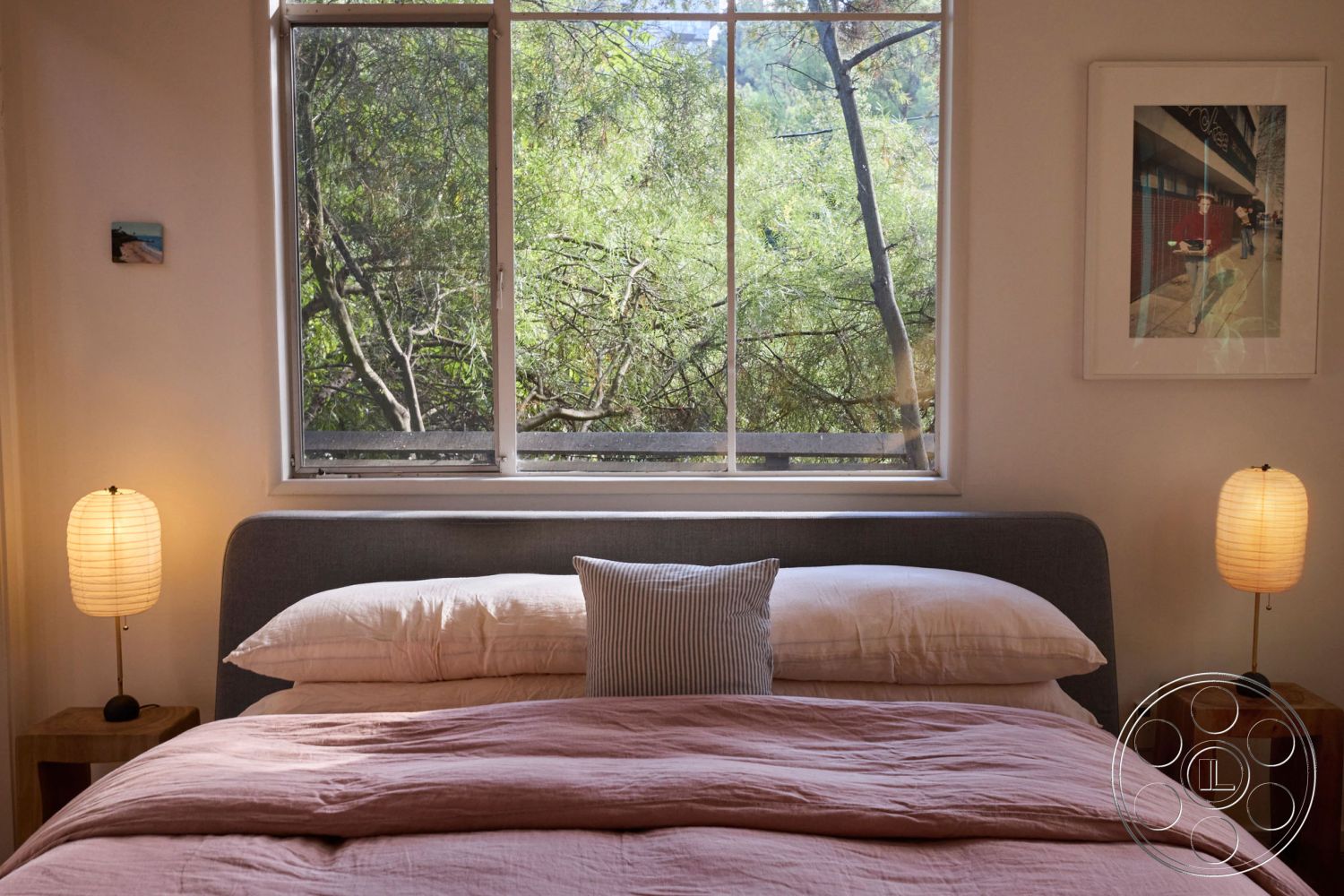 MCM 26 - earthy color palette, plush throw pillows, wooden nightstands, interior bedroom, white painted walls, natural light flooding in, soft pink bedding, hardwood floor bedroom, king-size bed with gray upholstered headboard, large double-pane window with forest view, minimalist bedside lamps, decorative art framed on wall, cozy and serene atmosphere, textured bedding fabric, modern Scandinavian design elements, greenery visible outside the window