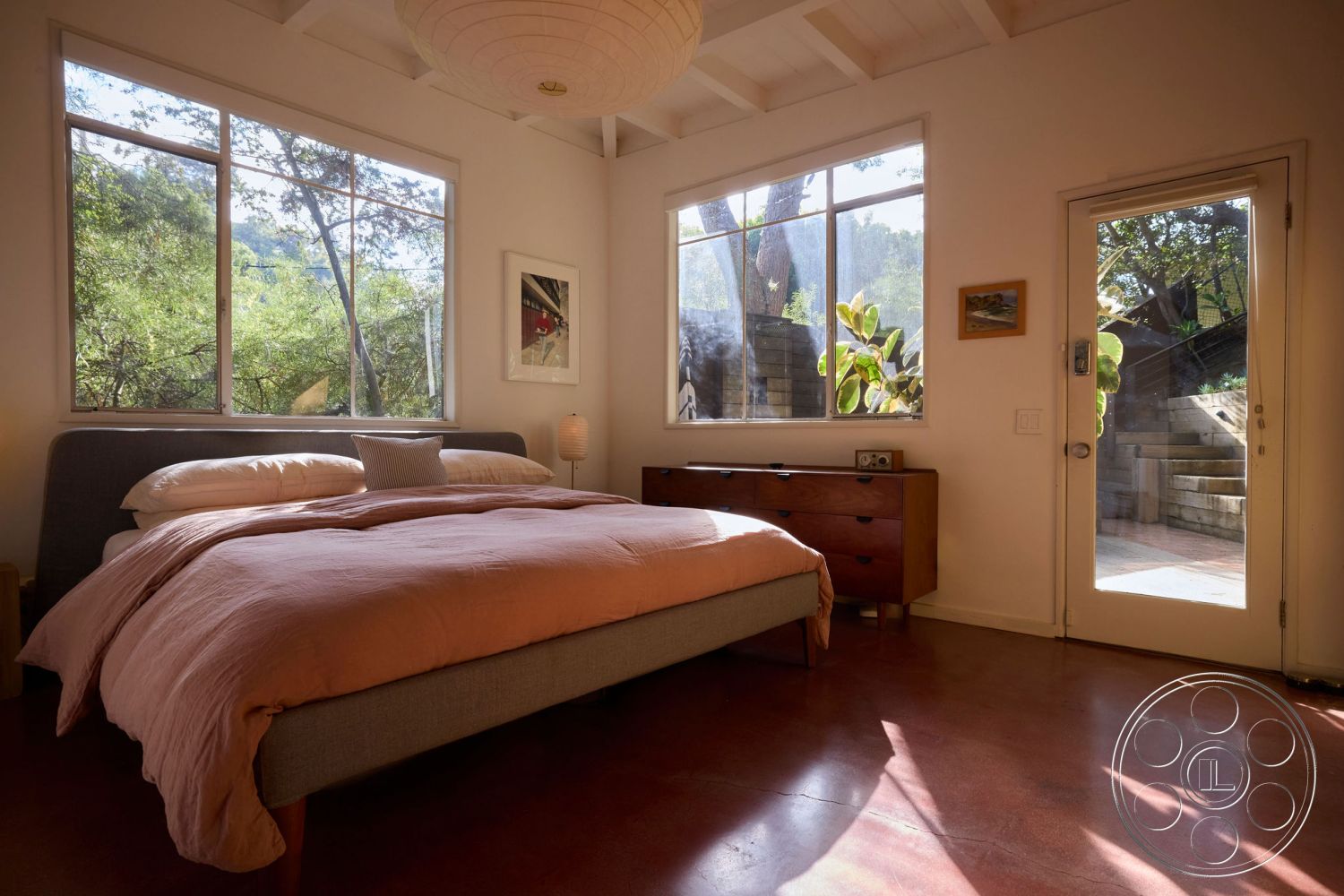 MCM 26 - minimalist decor, floor-to-ceiling windows, soft natural light, nature view through windows, wood beam ceiling, queen-sized upholstered bed, pink bedding set, mid-century modern dresser, polished concrete bedroom floors, plant-filled bedroom, simple artwork on walls, glass patio door