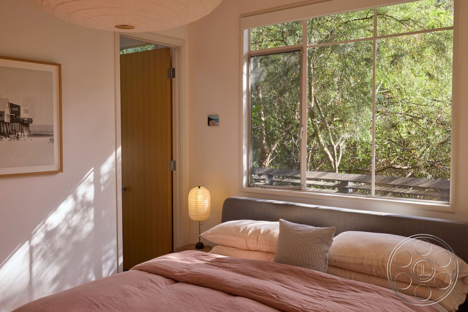 MCM 26 - minimalist decor, neutral color palette, natural light from large windows, modern pendant light fixture, interior bedroom, textured wall paint, soft pink bedding, wooden door with smooth finish, framed beach photograph on the wall, light wood trim around windows