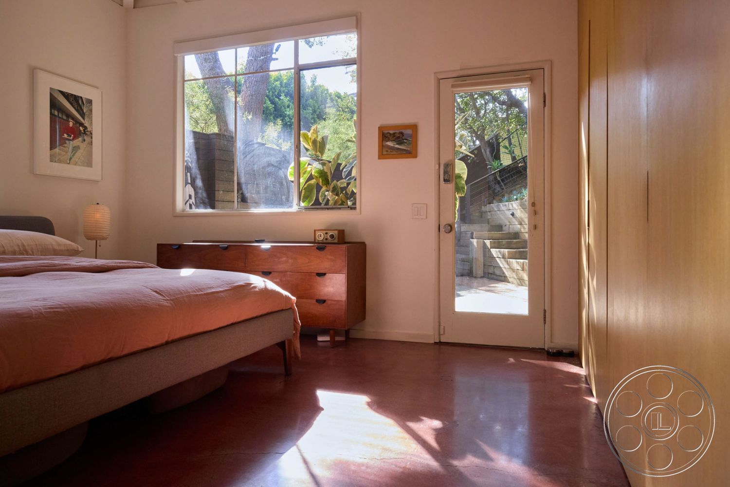 MCM 26 - natural light, minimalist design, cozy atmosphere, concrete floor, neutral wall color, wall-mounted art, outdoor view, floor-to-ceiling window, exposed wood ceiling beams, interior bedroom, large picture window, pink bedding set, red ceramic lamp, dark hardwood dresser, wooden sliding door, sunlit room
