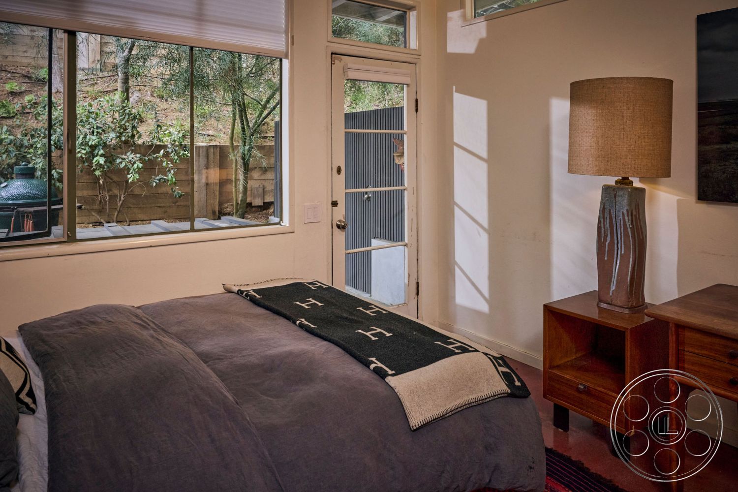 MCM 26 - neutral color palette, hardwood bedroom floors, minimalist bedside table, painted drywall walls, natural light-filled room, textured fabric throw blanket, interior bedroom with large rectangular windows, ceramic table lamp, modern art wall hanging, wooden framed ceiling, door leading to exterior garden patio, greenery visible from window, simple modern furnishings