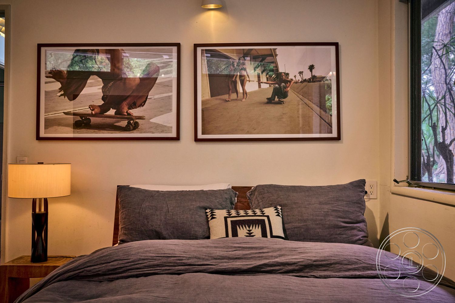 MCM 26 - neutral color palette, decorative throw pillow, textured wall surface, contemporary bedside lamp, cozy reading nook, wood bedside table, soft fabric textures, natural light interior, minimalist wall art, large window with nature view, interior bedroom decor, gray cotton bedding, framed art photographs, modern bedroom design, casual bohemian style, warm bedroom ambiance, pillow arrangement on bed