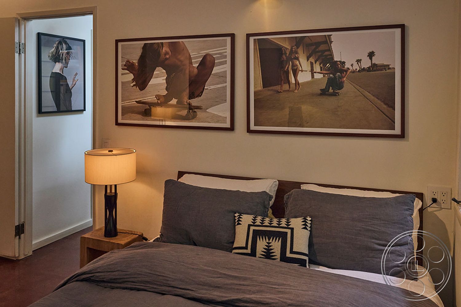 MCM 26 - neutral color palette, minimalist design elements, wooden bed frame, textured wall treatment, decorative throw pillows, soft lighting ambiance, interior bedroom space, concrete floor finish, functional space utilization, open doorway to hallway, gray duvet cover, fabric artwork decor, modern framed photographs, lamp with white fabric shade, nightstand with wooden finish, casual bedroom layout, natural light from hallway