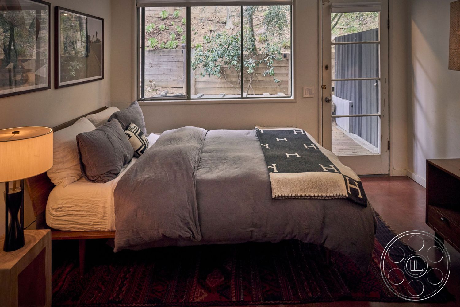 MCM 26 - wooden bed frame interior, neutral color palette interior, floor-to-ceiling windows interior, concrete flooring interior, sliding glass door interior, wooden nightstand interior, textured throw blanket interior, decorative pillows interior, glass door access interior, windows with natural light interior, artistic wall decor interior, natural wood panel wall interior, rugged textile area rug interior, modern bedside lamp interior, indoor plants view exterior, hillside view exterior, wooden privacy fence exterior