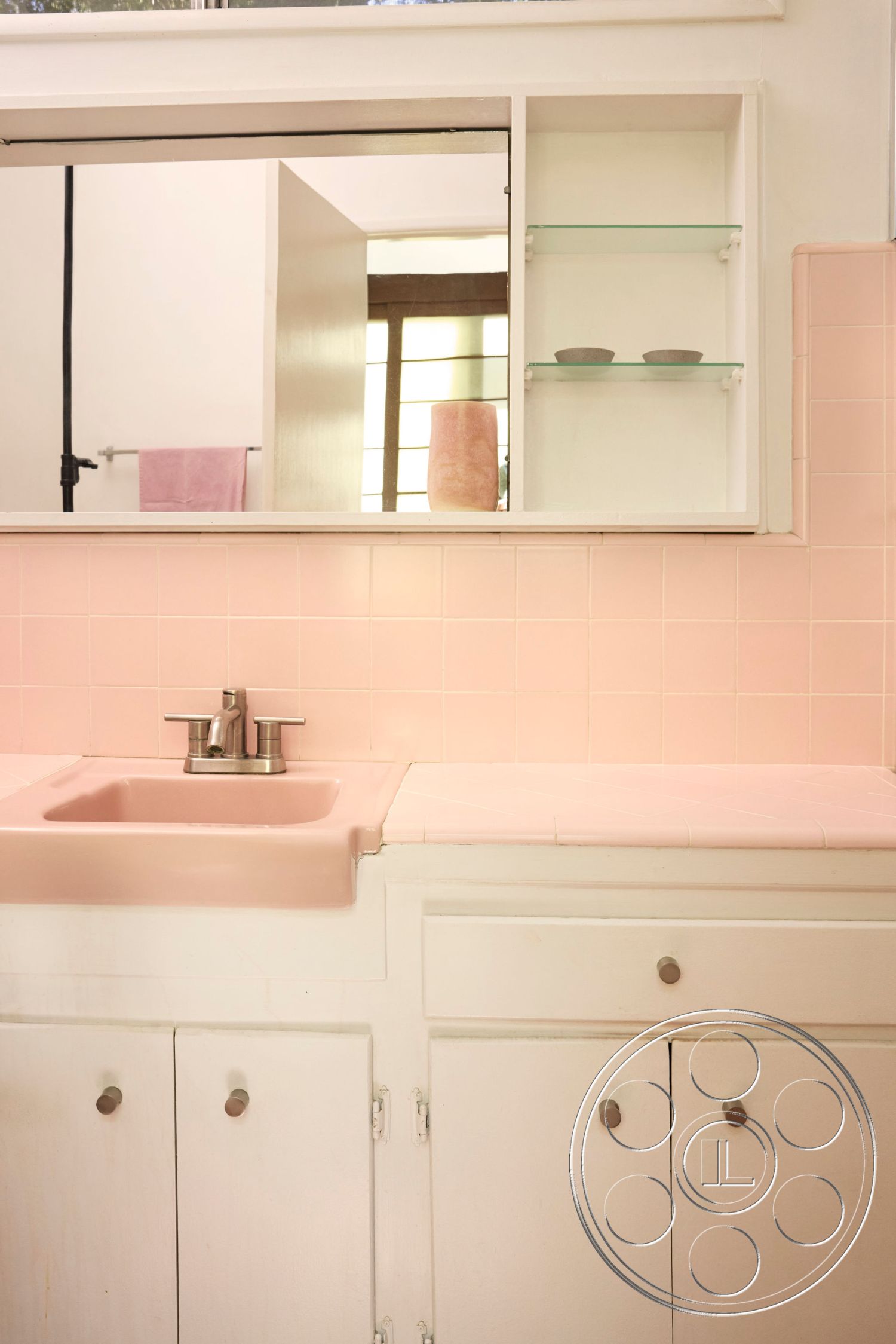 MCM 26 - interior natural light from window, interior pink tile bathroom walls, interior pink countertop with integrated sink, interior white cabinetry with silver knobs, interior mirror above sink, interior glass shelf storage, interior soft pink color scheme, interior modern design fixtures, interior tiled backsplash above countertop, interior minimalist bathroom style