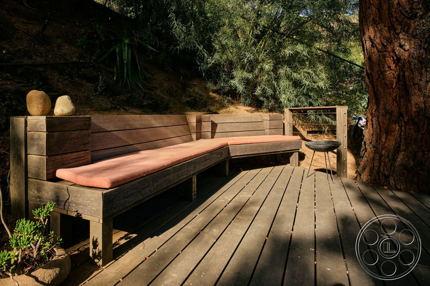 MCM 26 - modern landscape design, integrated landscaping, sun-drenched patio, earth-tone color palette, exterior wooden deck, lush greenery surrounding, natural stone decorative elements, L-shaped built-in bench seating, cushioned outdoor seating, weathered wood planks, rustic outdoor fire pit, textured wood railings, functional outdoor living space, private backyard setting, intimate gathering area, natural outdoor materials