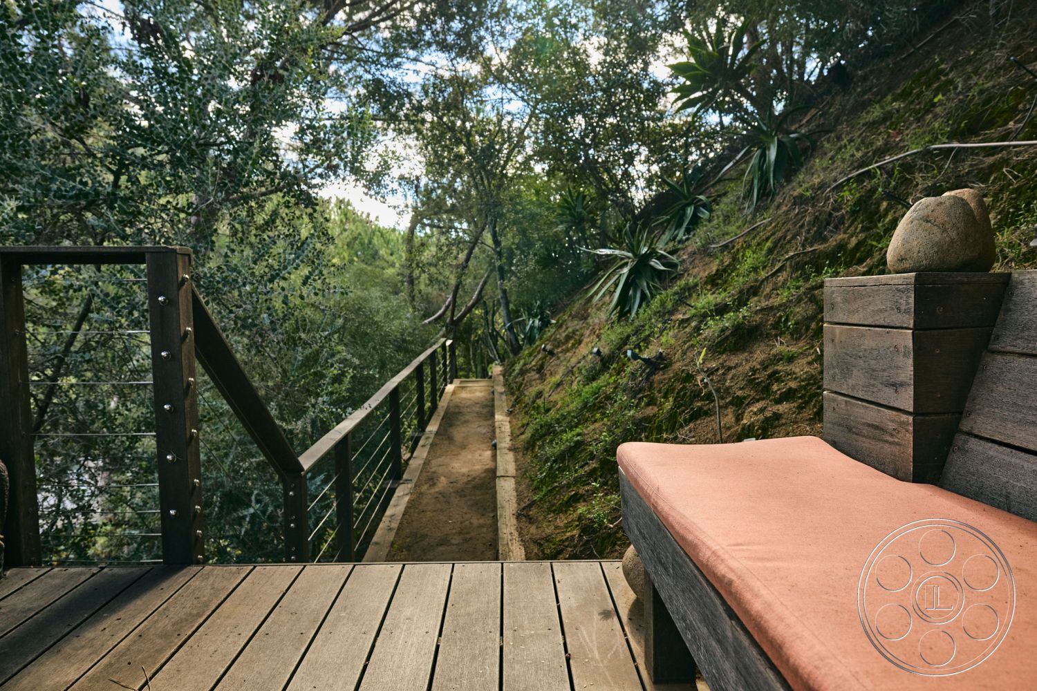 MCM 26 - exterior deck with wooden planks, stainless steel railing with cable detail, outdoor seating area with cushions, natural stone accent decor, surrounded by wooded landscape, pathway leading into forest, rustic wooden bench with backrest, sloped terrain with greenery, nature-inspired outdoor space, casual outdoor lounging area