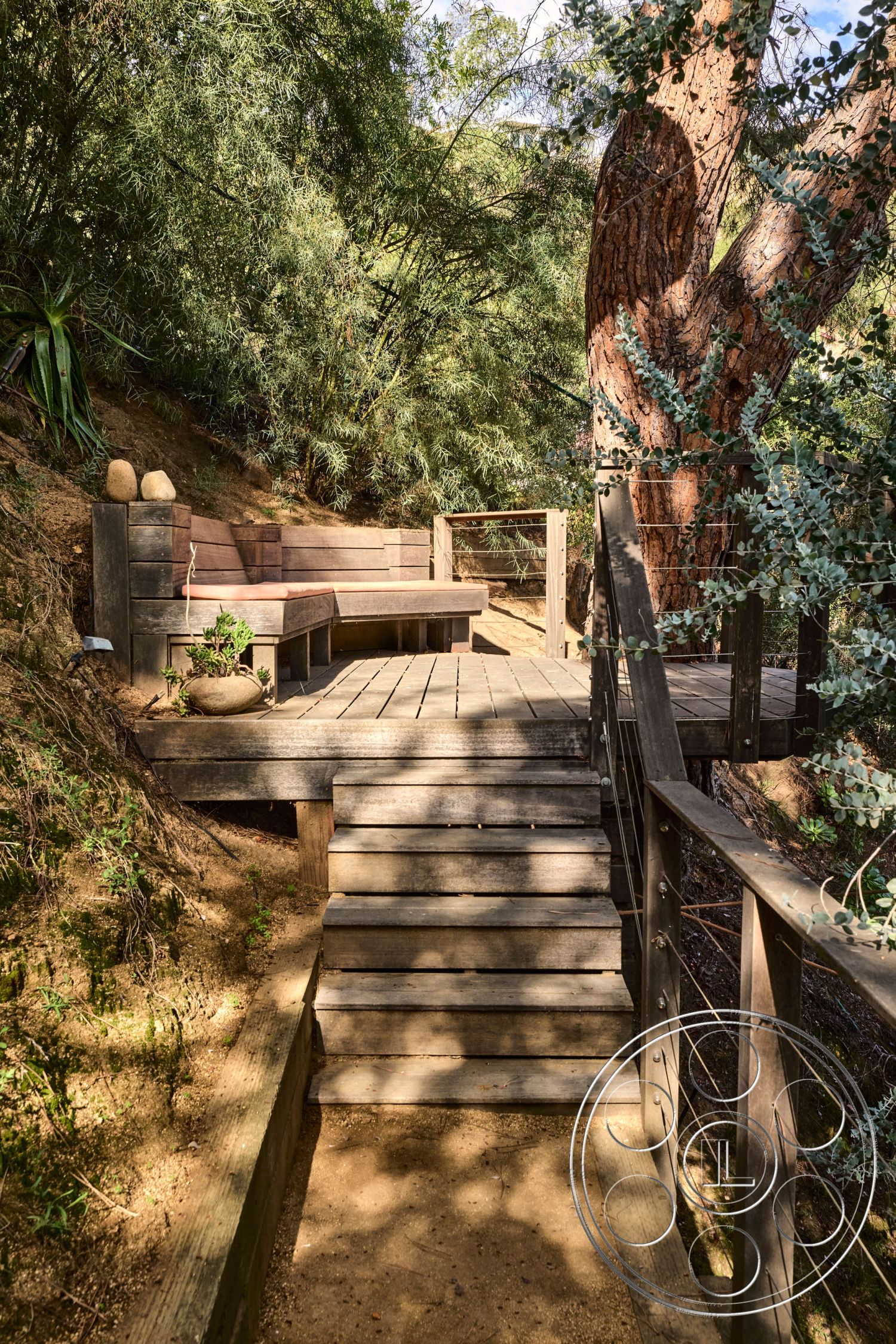 MCM 26 - natural landscape, surrounded by greenery, elevated platform, earth-toned color palette, wooden deck flooring, rustic outdoor setting, wood bench seating, interior outdoor space, metal cable railing, light-filtering shadows, stairs with wooden treads, textured wood details, slope terrain