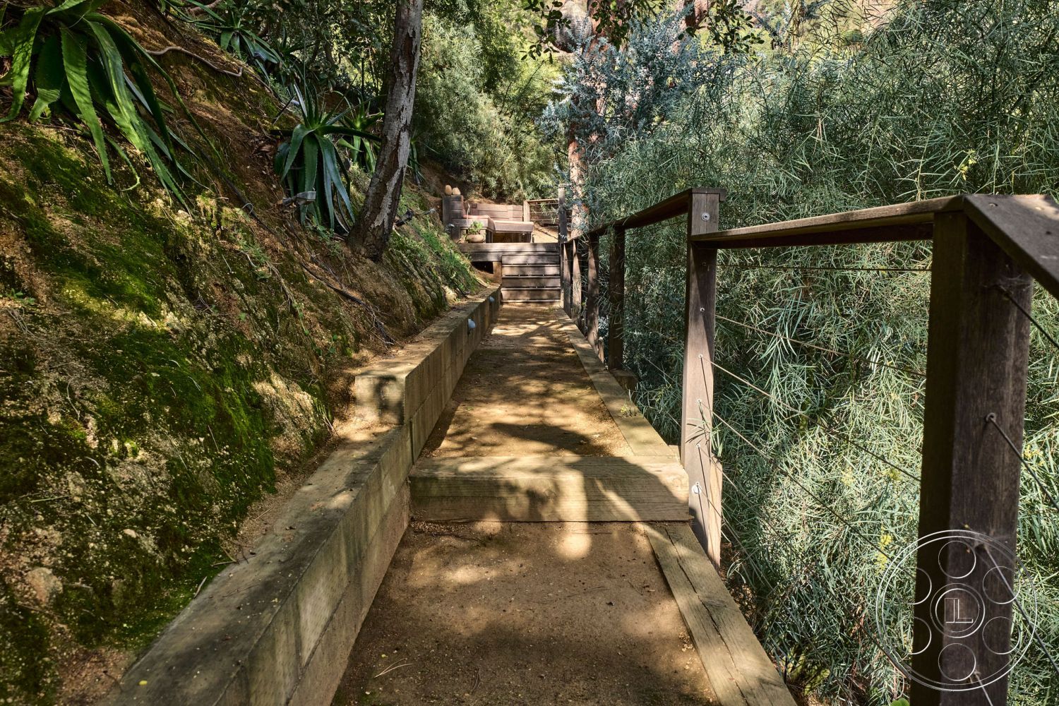 MCM 26 - soft natural lighting, surrounding lush greenery, natural stone border, environmentally integrated landscaping, wooden walkway path, natural earth landscaping, exposed wooden railing details, hillside path setting, integrated natural steps, outdoor trail design, rustic wooden structure, wooden park bench area, winding path shape, wood beam support structure, interior/exterior harmony