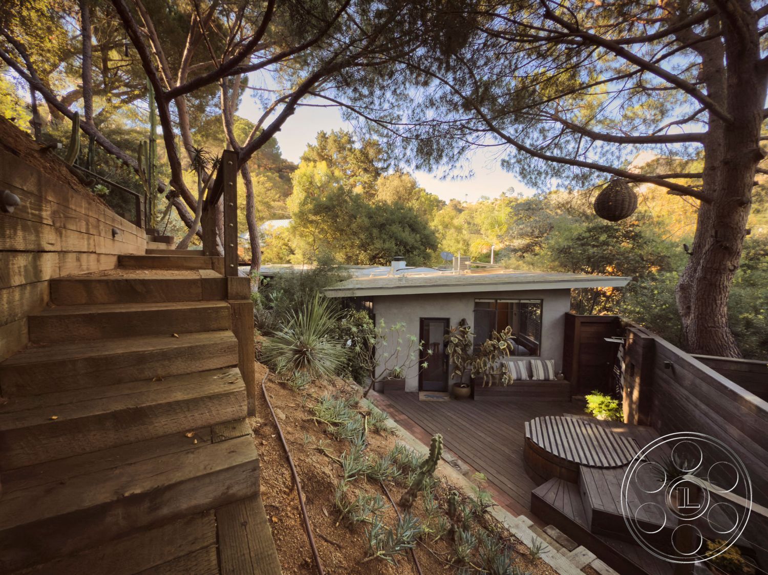 MCM 26 - floor-to-ceiling windows, decorative outdoor lighting, stucco exterior walls, interior minimalistic design, modern flat roof design, landscaped garden patio, native plant landscaping, modern outdoor living space, urban forest setting, exterior wooden railing, wooden staircase steps, wraparound wooden deck, round wooden seating area, textured natural hillside, tree canopy shade