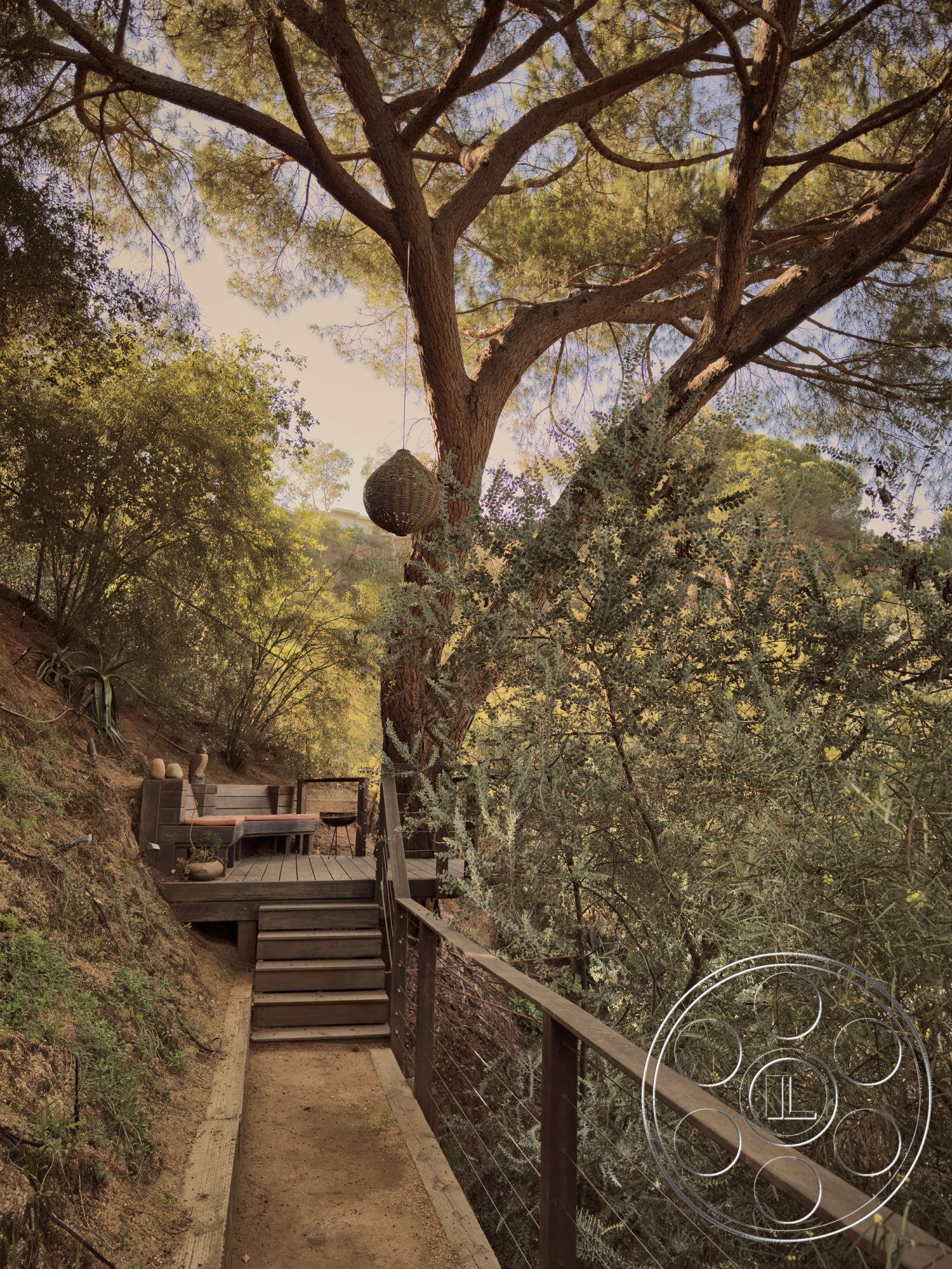 MCM 26 - natural materials, outdoor seating area, sloped terrain, interior-exterior flow, gravel pathway, surrounded by greenery, natural wood furniture, sustainable design elements, minimalist patio design, wood deck patio, landscape setting, wood railing details, tree canopy shade, rustic outdoor living space, tranquil nature retreat