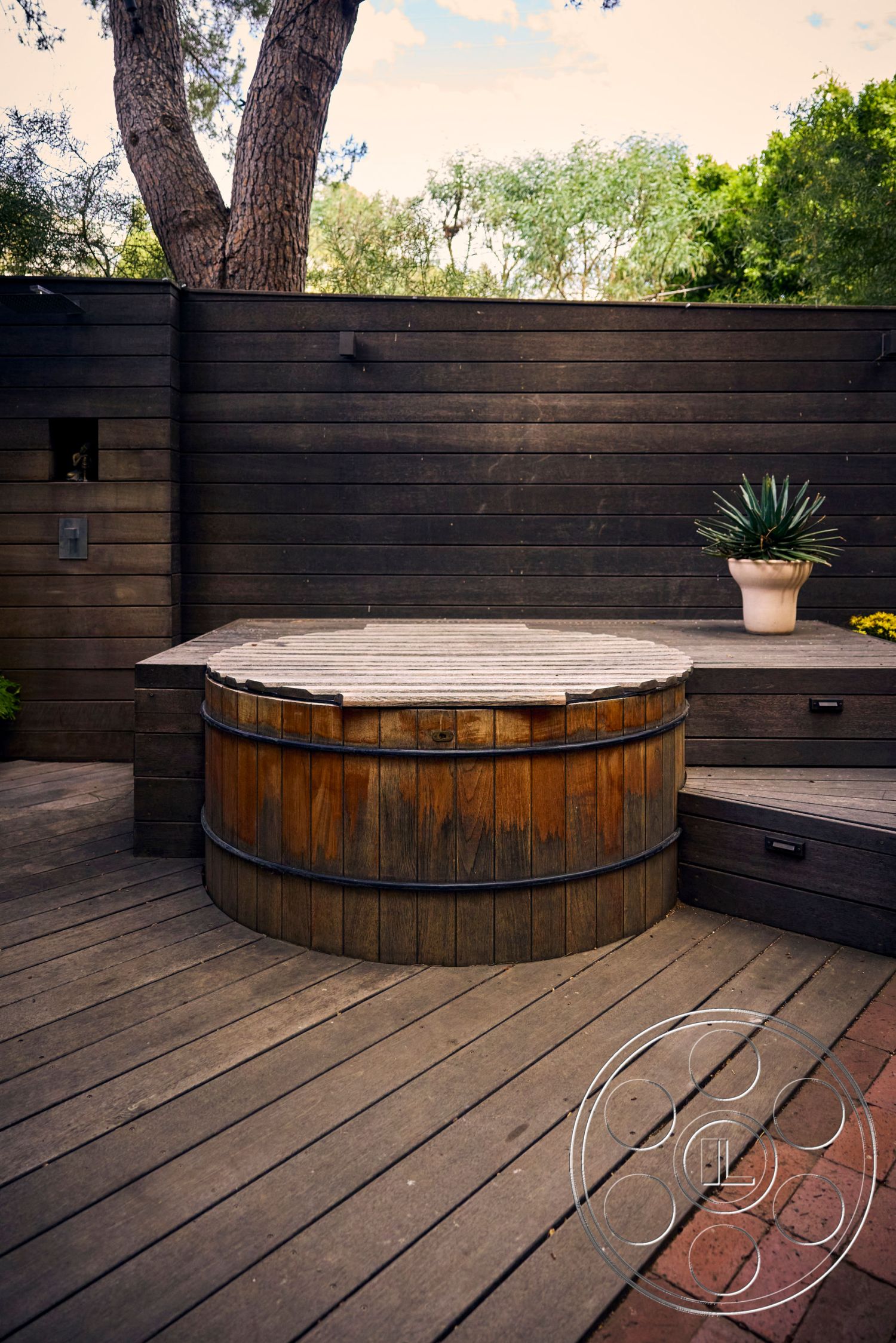 MCM 26 - surrounding greenery, outdoor living space, natural wood finishes, landscaped garden setting, exterior wooden deck flooring, textured wood surface, round wooden hot tub, rustic wooden barrel design, horizontal wooden slat privacy wall, multilevel deck design