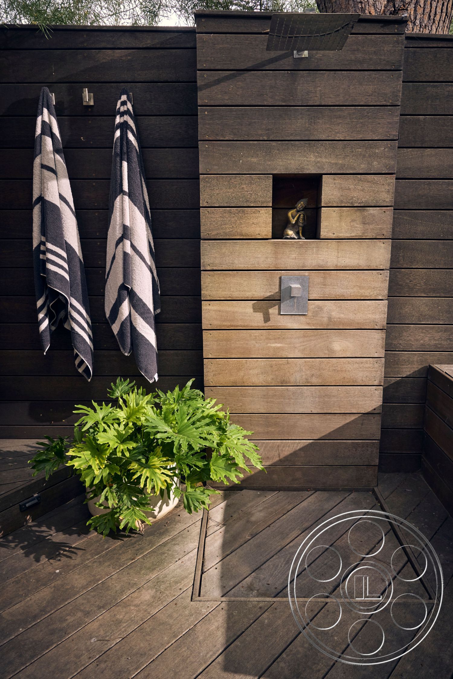 MCM 26 - surrounding greenery, open-air layout, modern minimalistic design, natural wood deck flooring, exterior wooden wall cladding, horizontal wood slat panels, built-in outdoor shower fixture, decorative stone niche with statue, contrasting black and white beach towels, potted tropical plant, angular outdoor layout, natural light and shadow interplay, private garden setting, cozy outdoor retreat