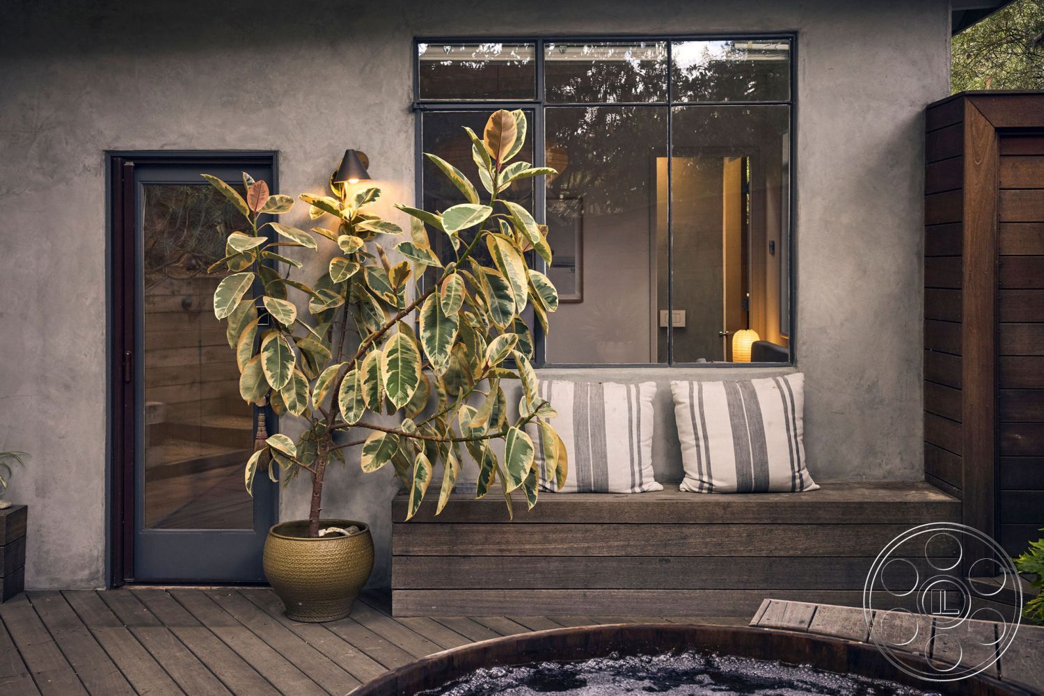 MCM 26 - earthy color palette, inviting entryway, relaxed seating area, cozy outdoor nook, functional outdoor space, striped outdoor cushions, natural wood accent wall, textured plaster wall, exterior wood deck flooring, exterior concrete wall, contemporary door design, wood plank seating area, oversized glass window, modern planter with foliage, minimalist outdoor setting, urban garden environment, sculptural plant arrangement, peaceful home exterior