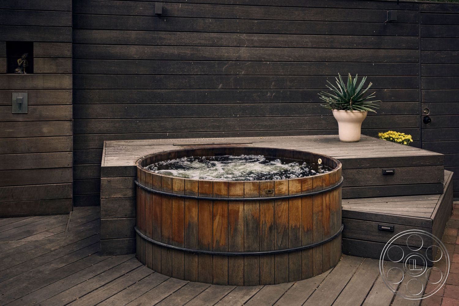 MCM 26 - minimalist design, relaxation space, dark wood deck, suburban backyard setting, exterior wooden hot tub, rounded wooden hot tub, textured wood paneling walls, concrete step risers, planter with agave plant, yellow flowering plant, contrast of wood and concrete materials, modern outdoor spa area, smooth wood surface