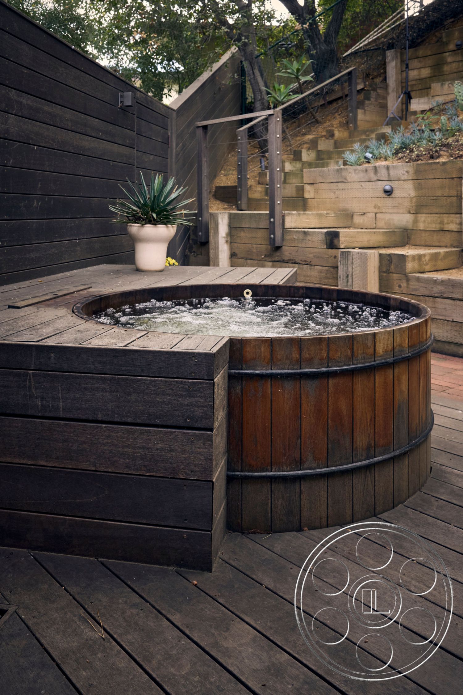 MCM 26 - natural materials, earthy color palette, natural wood deck, private outdoor retreat, landscaped hillside setting, composite deck flooring, wooden hot tub surround, exposed wooden wall panels, modern exterior stairs, staircase with cable railing, curved wood steps, outdoor garden area with planters, textured wooden step walls, interior/exterior integration, multilevel outdoor space, rustic hot tub design, sustainable wood elements