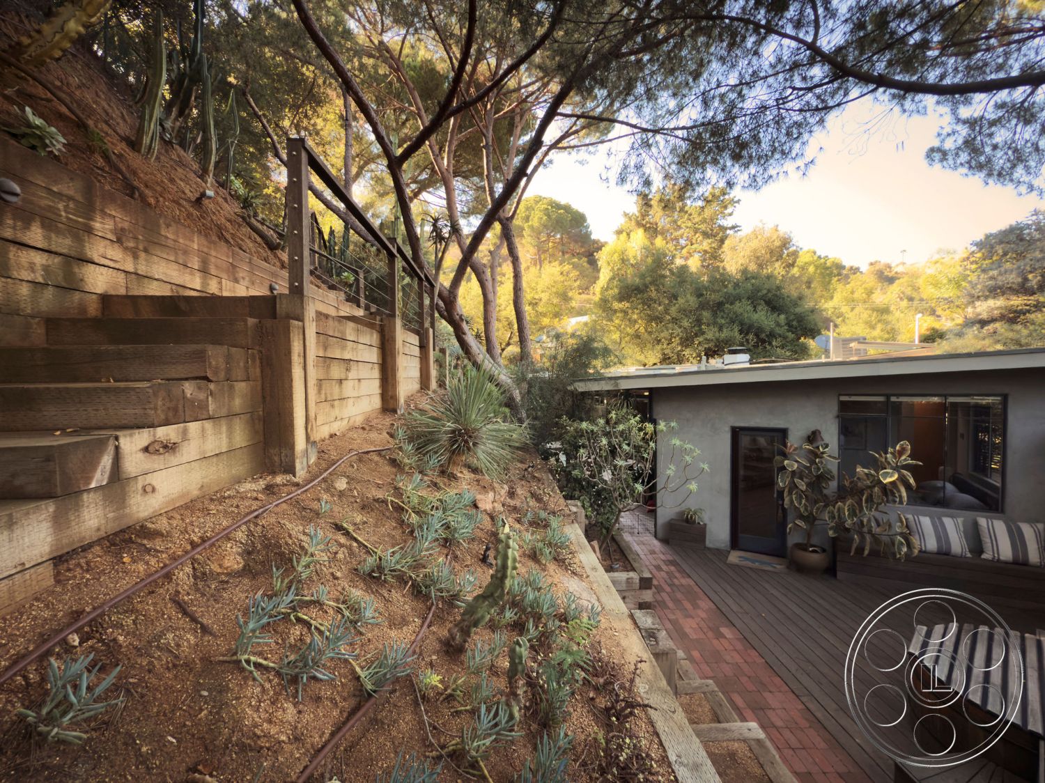 MCM 26 - earthy color palette, glass sliding doors, sustainable landscaping, concrete patio flooring, interior open layout, modern architectural details, integrated outdoor seating, landscaped hillside setting, minimalist home design, urban hillside property, exterior living space, wood retaining wall, wood staircase steps, natural landscape garden, wood deck surface, horizontal wood slats railing, surrounding forest view