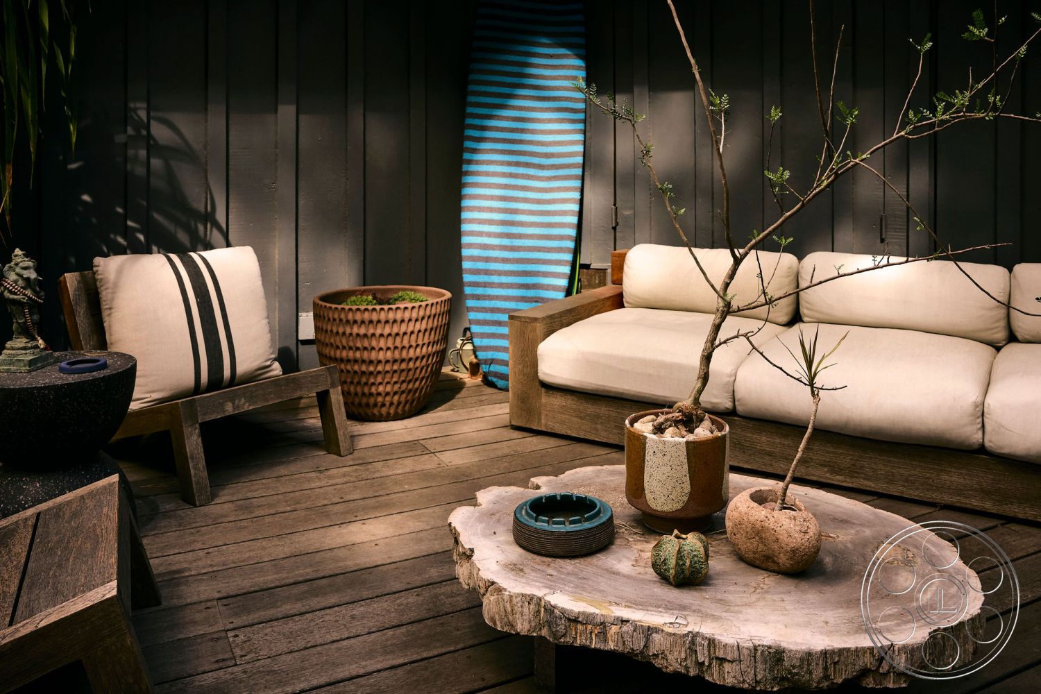 MCM 26 - wooden patio flooring, natural light-filled space, minimalist patio design, rustic outdoor seating area, textured stone planter, exposed wooden wall panels, outdoor sectional sofa with cream cushions, ceramic planter with textured finish, natural wood coffee table slab, striped outdoor throw blanket, dark wooden lounge chair with slatted design, modern outdoor décor accents, eco-friendly garden elements, potted plant with succulents, interior outdoor living area, cozy outdoor relaxation space