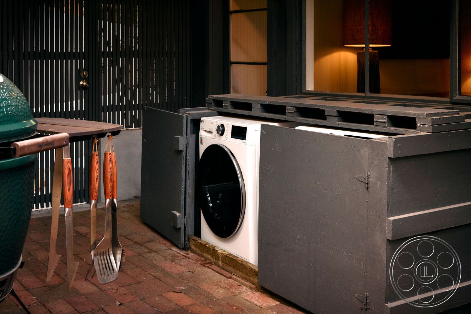 MCM 26 - wooden countertop, modern rustic design, functional outdoor storage, brick patio flooring, urban backyard setting, exterior laundry area, metal grill station, black wooden storage cabinet, white front-loading washer, stainless steel cooking utensils, slat privacy screen, outdoor kitchen space, available outdoor lighting, multi-functional outdoor space