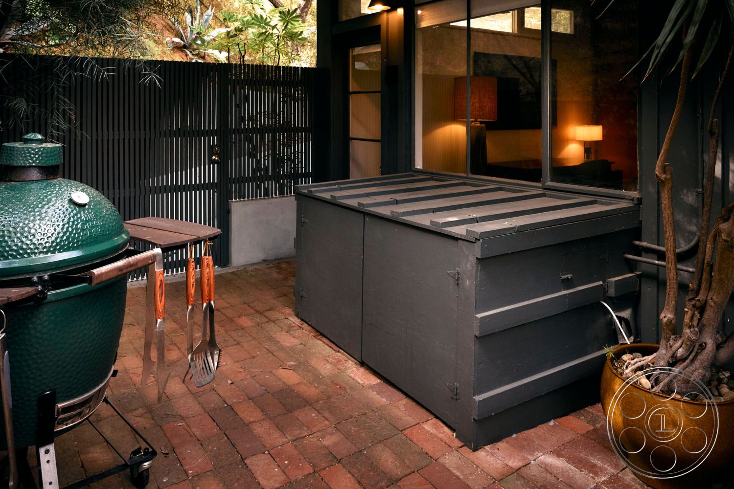 MCM 26 - textured wall treatments, dim ambient lighting, urban backyard, exterior patio area, brick patio flooring, modern fence design, tropical plant decor, green ceramic grill, contemporary outdoor setting, wooden grilling utensils, wood storage box with slanted lid, metal garden planter, glass sliding door leading to interior