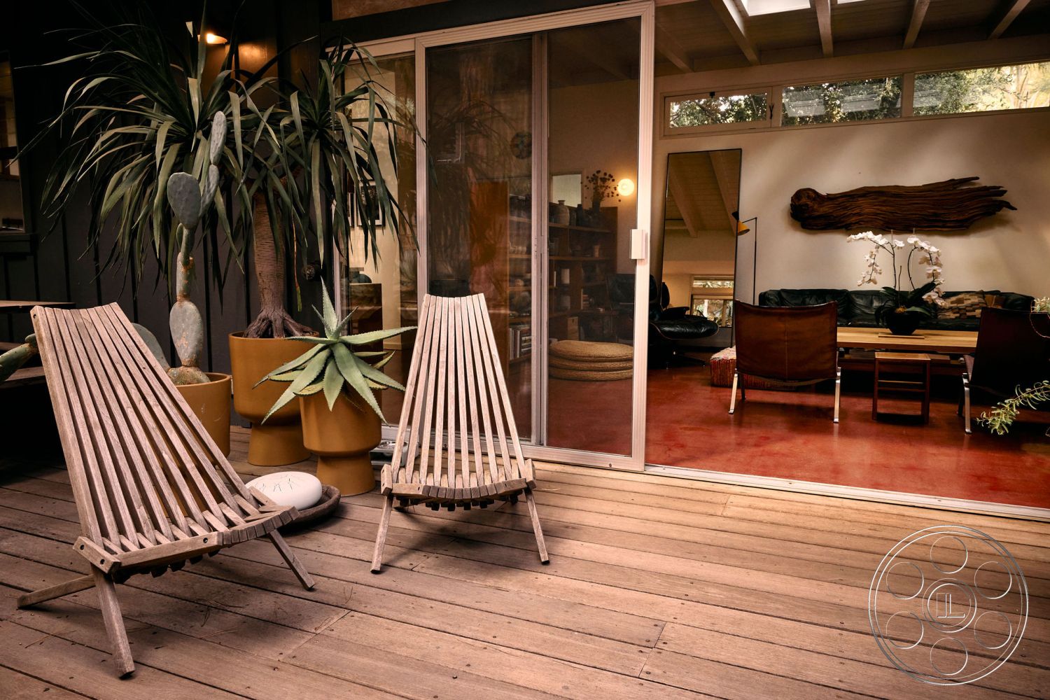 MCM 26 - textured wall accents, open concept living space, rustic wooden planters, contemporary aesthetic, wood beam ceiling, surrounding lush greenery, exterior wooden deck flooring, natural light from floor-to-ceiling windows, minimalist outdoor seating area, interior stained concrete flooring, modern wooden lounge chairs, potted desert plants, sliding glass door entrance, organic wooden decor pieces, bohemian style decor elements, casual outdoor patio atmosphere