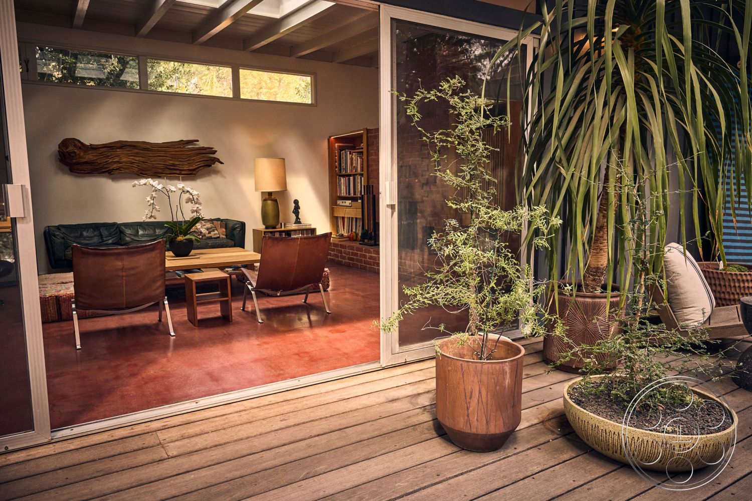 MCM 26 - wood coffee table, mid-century modern decor, wooden lounge chairs, exposed wood ceiling beams, natural light through large windows, potted indoor plants, modern leather sofa, glass sliding doors leading to patio, interior living room with concrete floor, decorative wood wall art, lampshade with warm light, textured wall with organic shapes, plant pots with natural finishes, rustic charm accents in furniture, cozy and inviting space