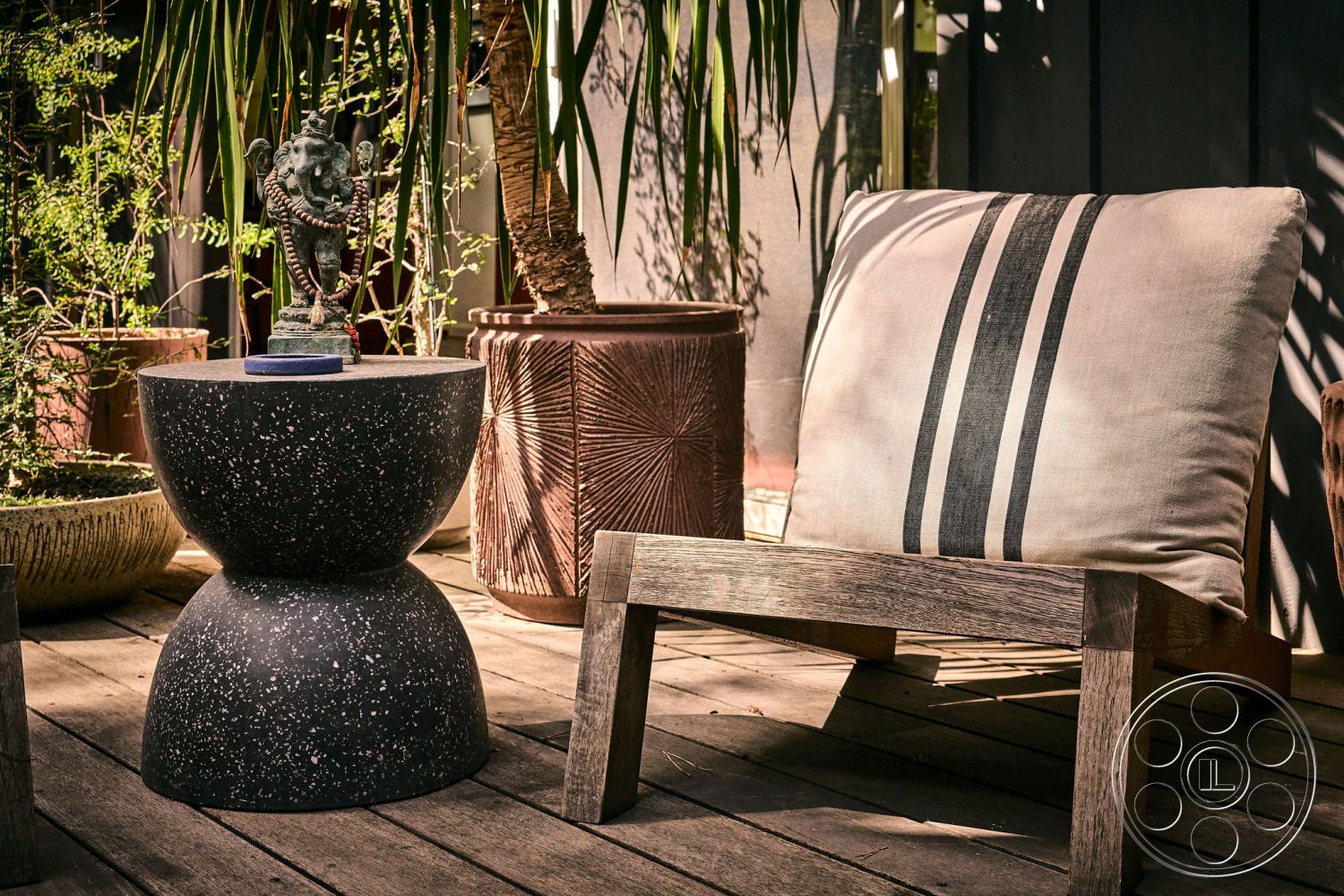 MCM 26 - decorative potted plants, wooden deck flooring, sculptural garden statue, natural light environment, exterior patio furniture, terrazzo side table, textured ceramic plant pots, cushioned lounge chair, striped outdoor pillow, tropical garden setting
