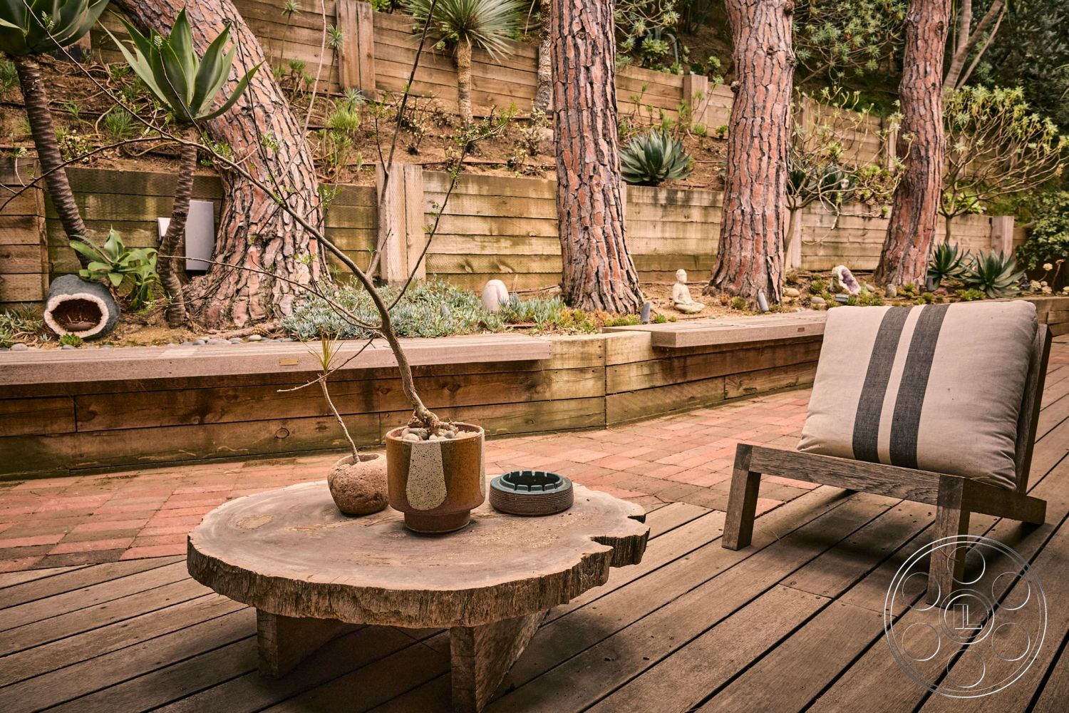 MCM 26 - contemporary outdoor seating, minimalist outdoor decor, landscaped garden setting, exterior wood decking, brick patio flooring, natural stone garden accents, private backyard retreat, natural wood outdoor table, textured wooden retaining wall, potted succulent arrangement, rustic log side table, lush green foliage backdrop, earth-toned ceramic planter, varied tree trunk textures, natural light outdoor space