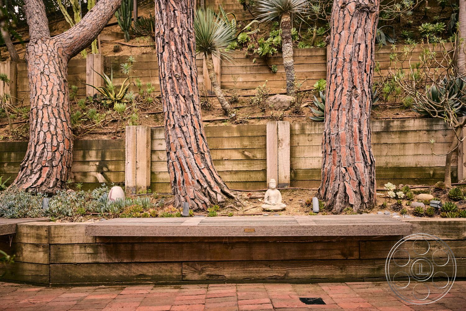 MCM 26 - exterior, earthy color palette, landscaped hillside, urban garden design, natural stone garden accents, succulent garden plants, red brick patio flooring, wooden retaining wall, exposed wooden bench seating, tall coniferous trees, meditative stone figure, desert planting environment, textured bark surface, secluded outdoor seating area