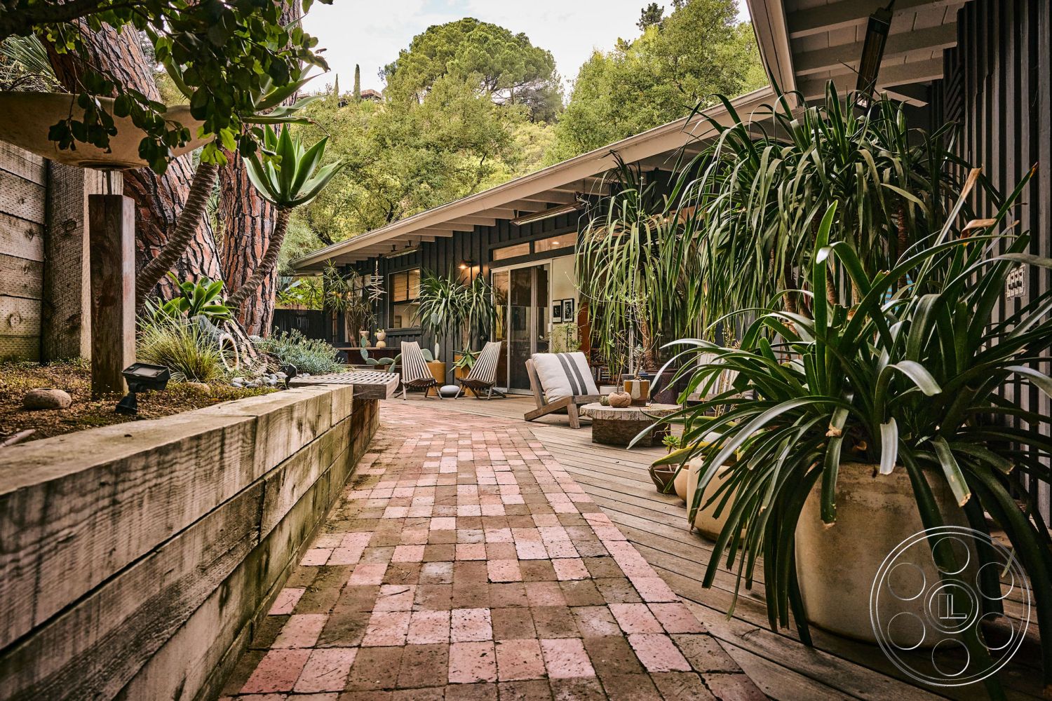 MCM 26 - contemporary outdoor seating area, outdoor patio area with lounge chairs, wood deck flooring exterior, sloped hillside setting, urban garden environment, brick pathway exterior, exposed wood beam structure exterior, lush landscaping with potted plants, modern sliding glass doors, textured concrete retaining wall exterior, flat roof design exterior, natural wood siding exterior, decorative stones in garden bed, open-air living space exterior, indoor-outdoor flow with large windows