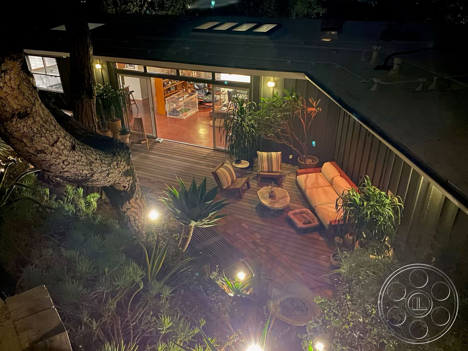 MCM 26 - large glass sliding doors, wooden coffee table, contemporary outdoor furniture, decorative stone planters, urban outdoor retreat, modern outdoor seating area, wood deck patio flooring, integrated indoor-outdoor living, relaxed outdoor living space, exterior lighting fixtures, natural wood beam ceiling, surrounding greenery landscaping, textured planter pots, interior open floor plan view, accent wall with plants, nighttime exterior ambiance, surrounding tree as natural feature, rustic natural wood furniture, cozy evening atmosphere, modern residence with outdoor connection