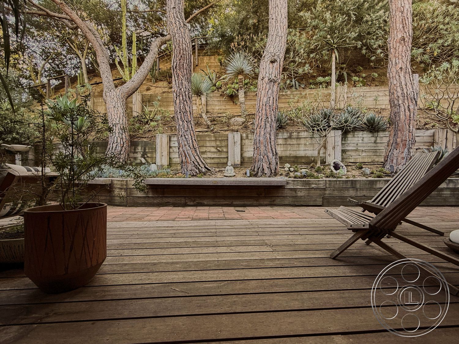 MCM 26 - interior garden view, outdoor lounge chairs, outdoor relaxation area, vertical wooden fence, landscaped garden setting, wooden deck flooring, rustic outdoor decor, exposed wood beam ceiling, natural light filtering through trees, tropical plant surroundings, multi-level garden design, red clay patio tiles, sloping terrain backdrop, planter pots with succulents, natural stone garden features, textured wood furniture, eco-friendly garden landscaping, serene outdoor atmosphere