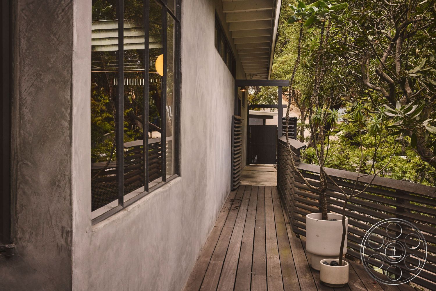 MCM 26 - floor-to-ceiling windows, contemporary architecture, textured plaster wall, minimalist architectural style, exposed wooden ceiling beams, exterior lighting fixtures, natural landscape backdrop, black metal railing details, outdoor planters with greenery, exposed concrete wall exterior, wood plank walkway, surrounding lush greenery setting, modern home entryway, concrete pathway leading to gate, interior natural light entry, wood overhang for shade, private garden area, sliding gate access