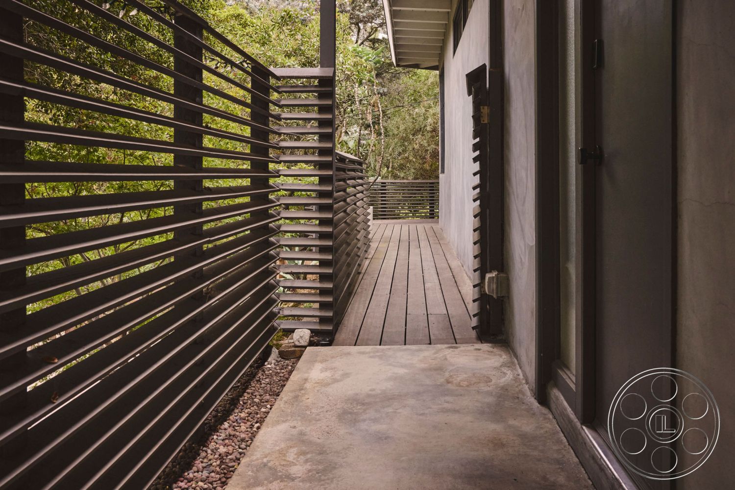 MCM 26 - textured concrete surface, minimalist outdoor space, exterior concrete walkway, contemporary architectural design, surrounded by lush greenery, clean lines and geometric shapes, modern home facade, horizontal wood slat fencing, private outdoor corridor, wood decking pathway, natural stone gravel border, flush entrance door, smooth plaster exterior walls, natural light filtering through, tranquil setting amidst trees