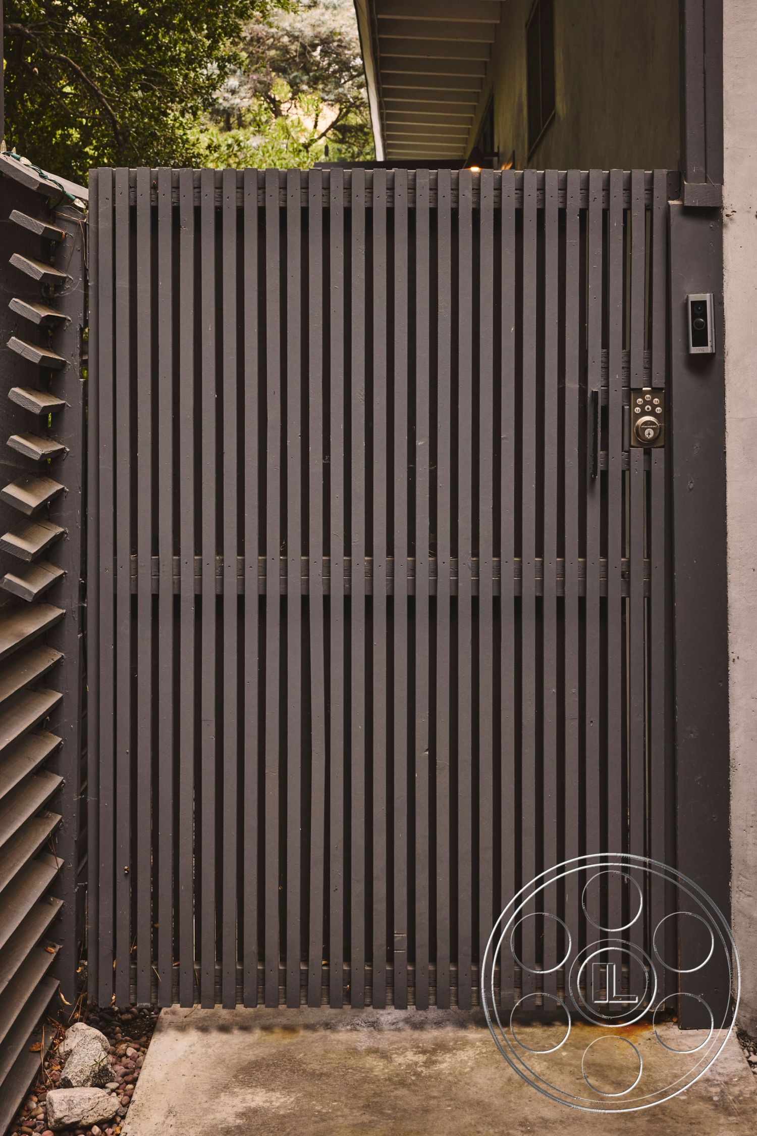 MCM 26 - textured concrete surface, natural greenery backdrop, modern architectural design, low-maintenance outdoor setting, landscaped garden surroundings, concrete pathway leading to gate, exterior slatted wooden gate, dark-stained vertical wood planks, integrated electronic keypad entry, minimalist fence design