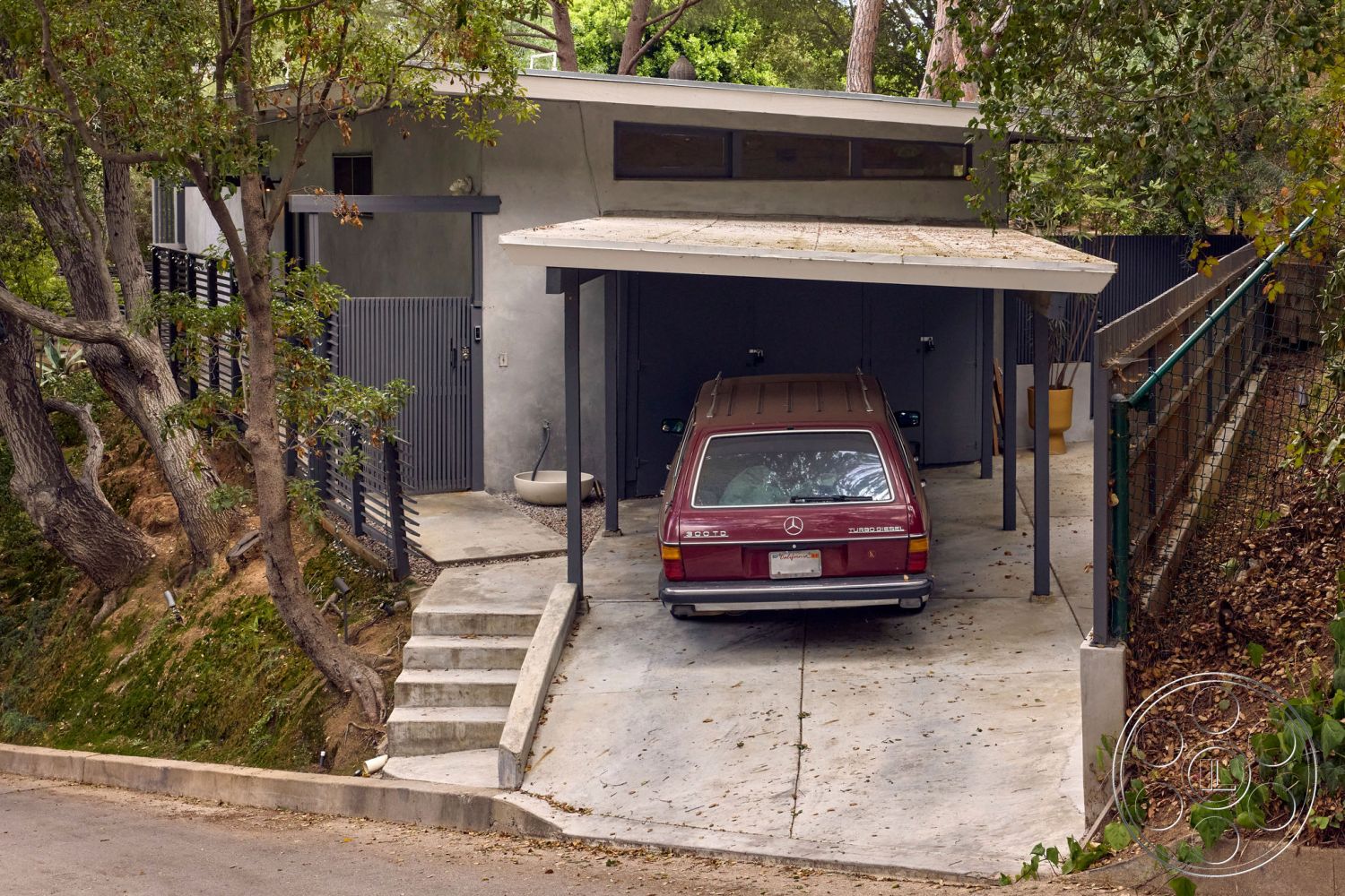 MCM 26 - large windows, exterior, contemporary design, natural landscaping, sloped terrain, surrounding trees, concrete steps, steel railing, modern architectural style, concrete driveway, wooden fence, dirt pathway, integrated outdoor space, outdoor lighting fixtures, urban hillside setting, open carport, minimalist entryway, flat roof carport, gray painted exterior walls