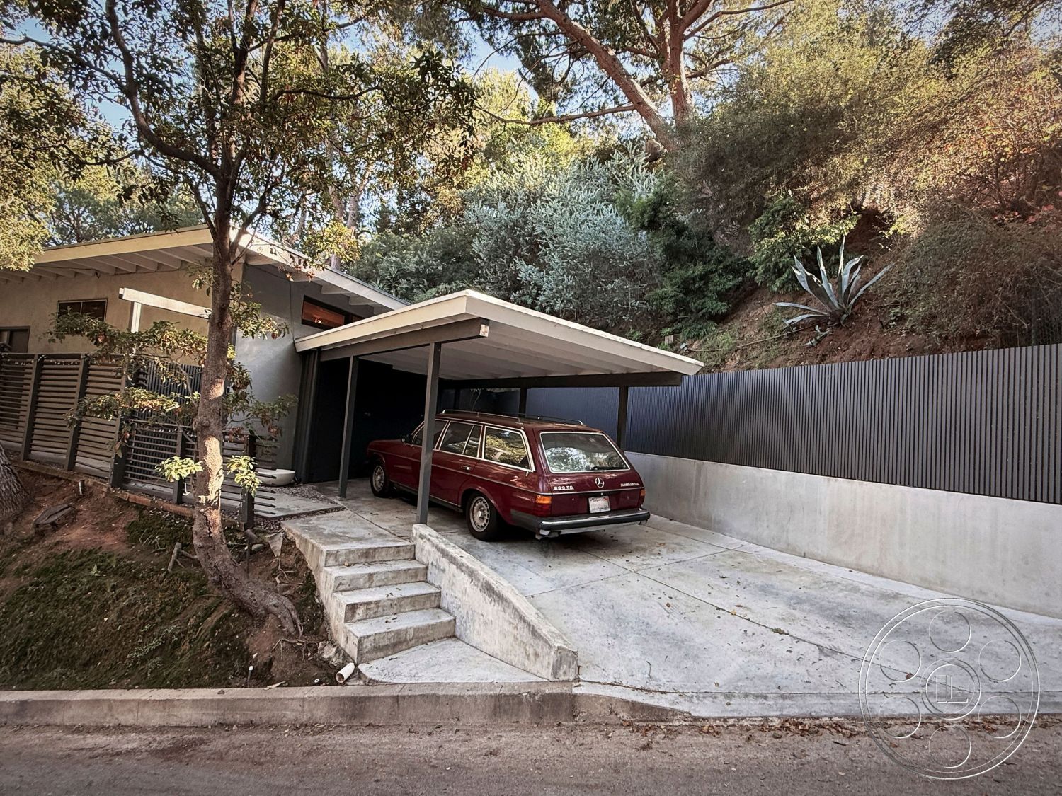 MCM 26 - exterior concrete driveway, exterior wood beam overhang, exterior minimalist architectural style, exterior natural vegetation, exterior concrete parking pad, exterior cantilevered roof structure, exterior steel railing, exterior vertical metal paneling, exterior landscaped hillside, exterior stone retaining wall, exterior sloped lot, exterior modern carport, exterior textured plaster wall