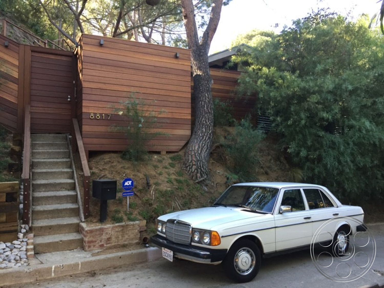 MCM 26 - natural stone accents, minimalist design elements, concrete steps, stepped pathway, mid-century modern architectural style, natural light emphasis, lush greenery surrounding, landscaped hillside setting, angular roof forms, exterior wood siding, horizontal wooden boards, raised deck area, built-in mailbox, wooden railing details, exposed aggregate concrete footing, sloped lot, ornamental shrubbery, off-street parking area, dual-tone wood texture, pine tree landscape