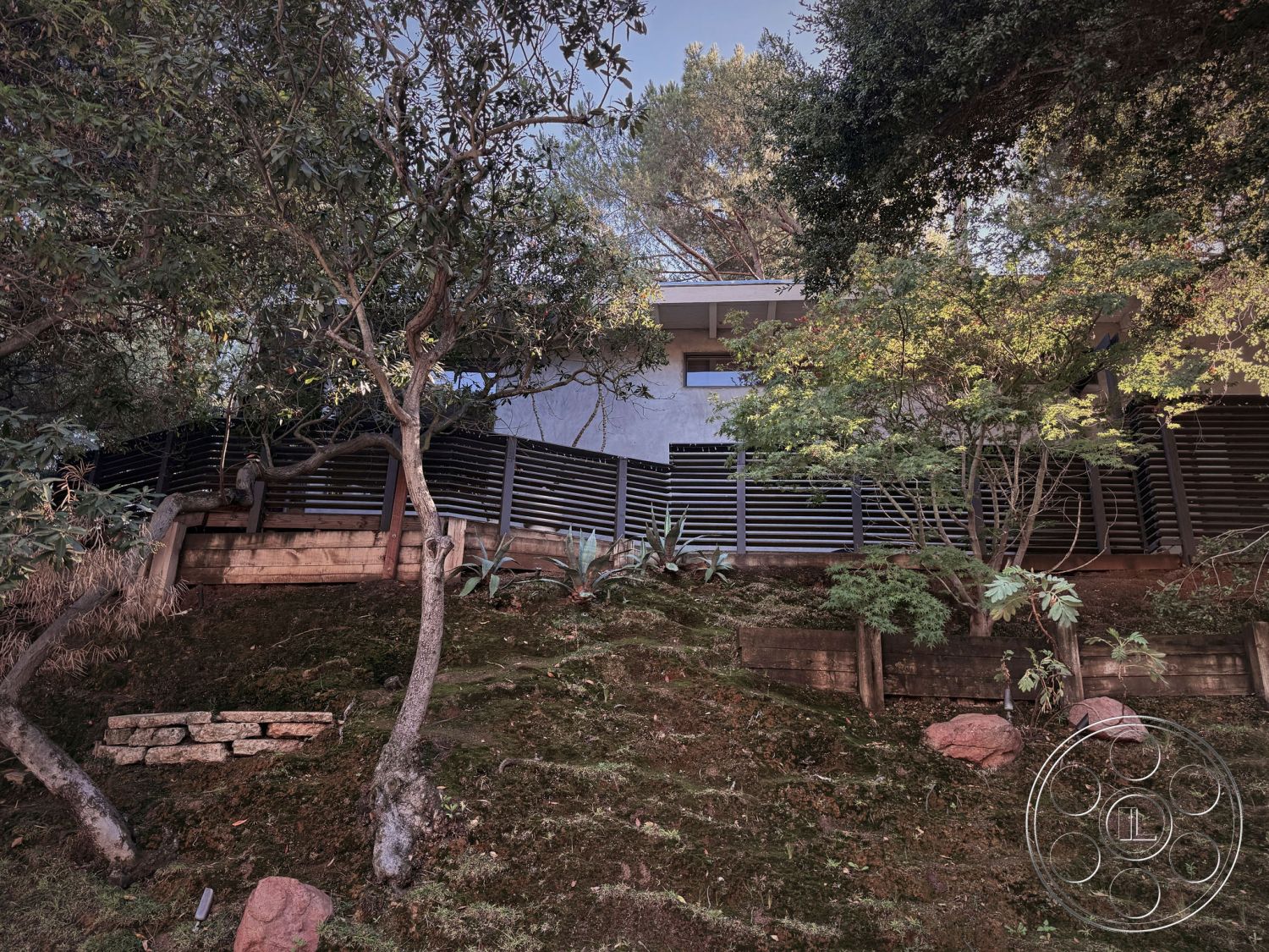 MCM 26 - exterior, open floor plan, large glass windows, modern architectural style, asphalt driveway, natural stone retaining wall, urban hillside location, wooden deck area, native plant landscaping, landscaped hillside garden, concrete foundation walls, wooden slat fencing, textured concrete exterior walls, minimalist roof design, sloped terrain setting