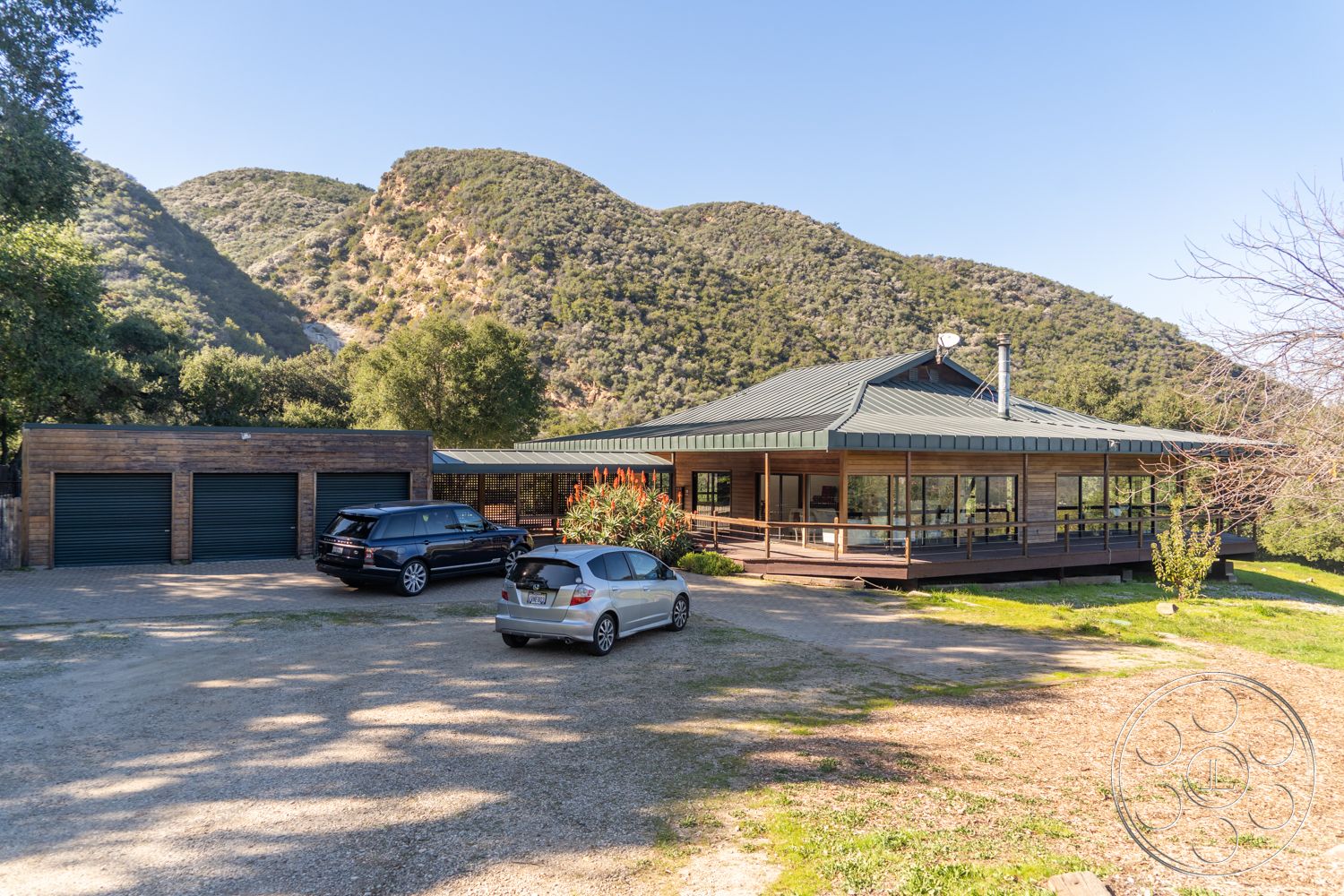 Topanga Retreat - natural stone accents, outdoor seating area, landscaped yard, exposed wood beams, floor-to-ceiling windows, concrete driveway, two-car garage, spacious lawn, gravel path, architectural detail, expansive outdoor space, wooden railings, terraced landscaping, sunlit exterior, green metal roof, mountain view setting, modern ranch-style home, rectangular patio, exterior wood siding, large deck area