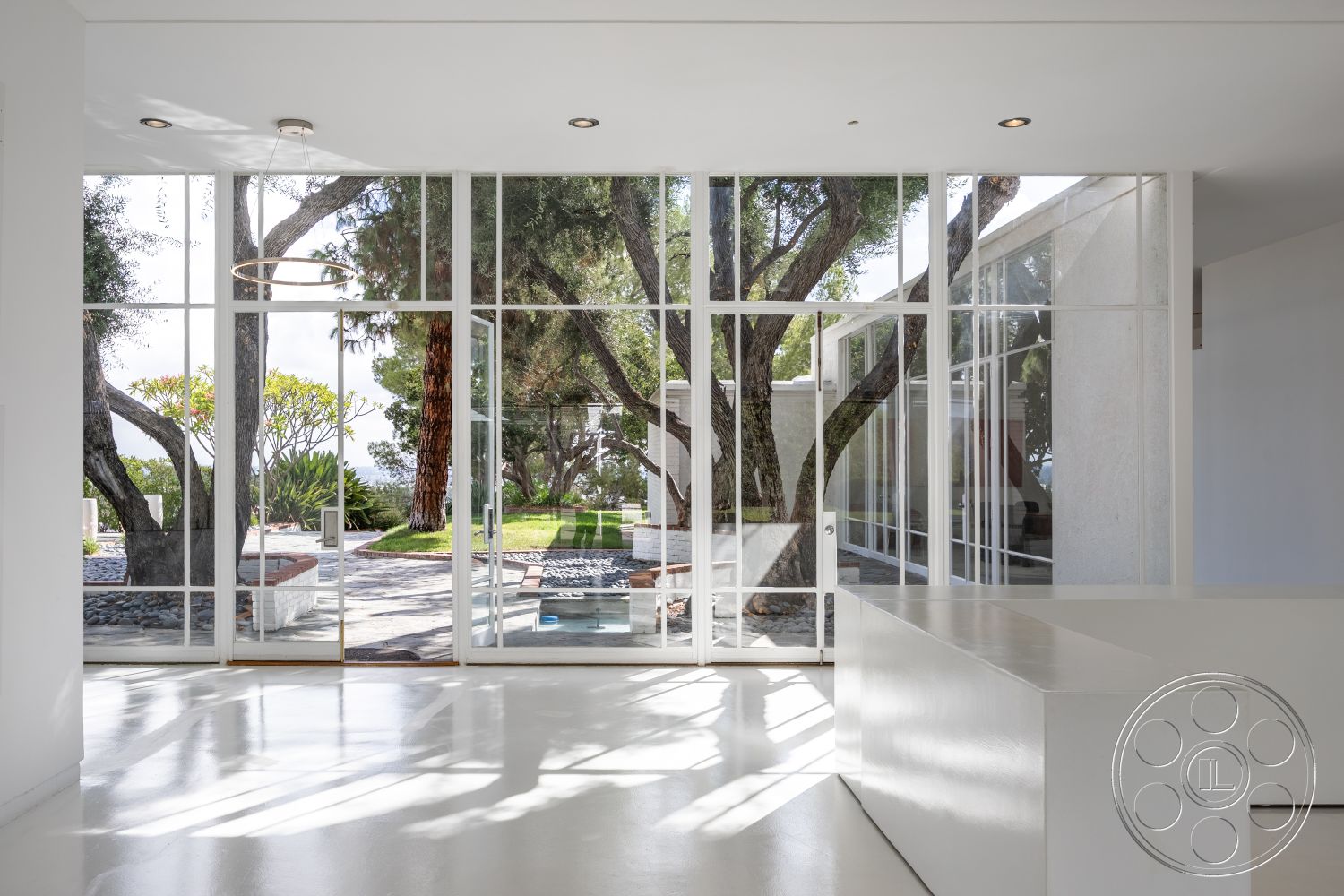 Sinatra House - floor-to-ceiling windows, seamless indoor-outdoor flow, contemporary lighting fixtures, modern minimalist design, white painted walls, open concept living area, interior glass partition walls, geometric shapes in architecture, city skyline view from large windows, exterior landscaped yard, concrete floor with polished finish, exposed wooden beams in ceiling, natural light indoor space, energy-efficient glass doors, landscaped outdoor area with mature trees, patio with stone and pebble landscaping, sleek white kitchen island, modern aesthetic home, functional indoor space