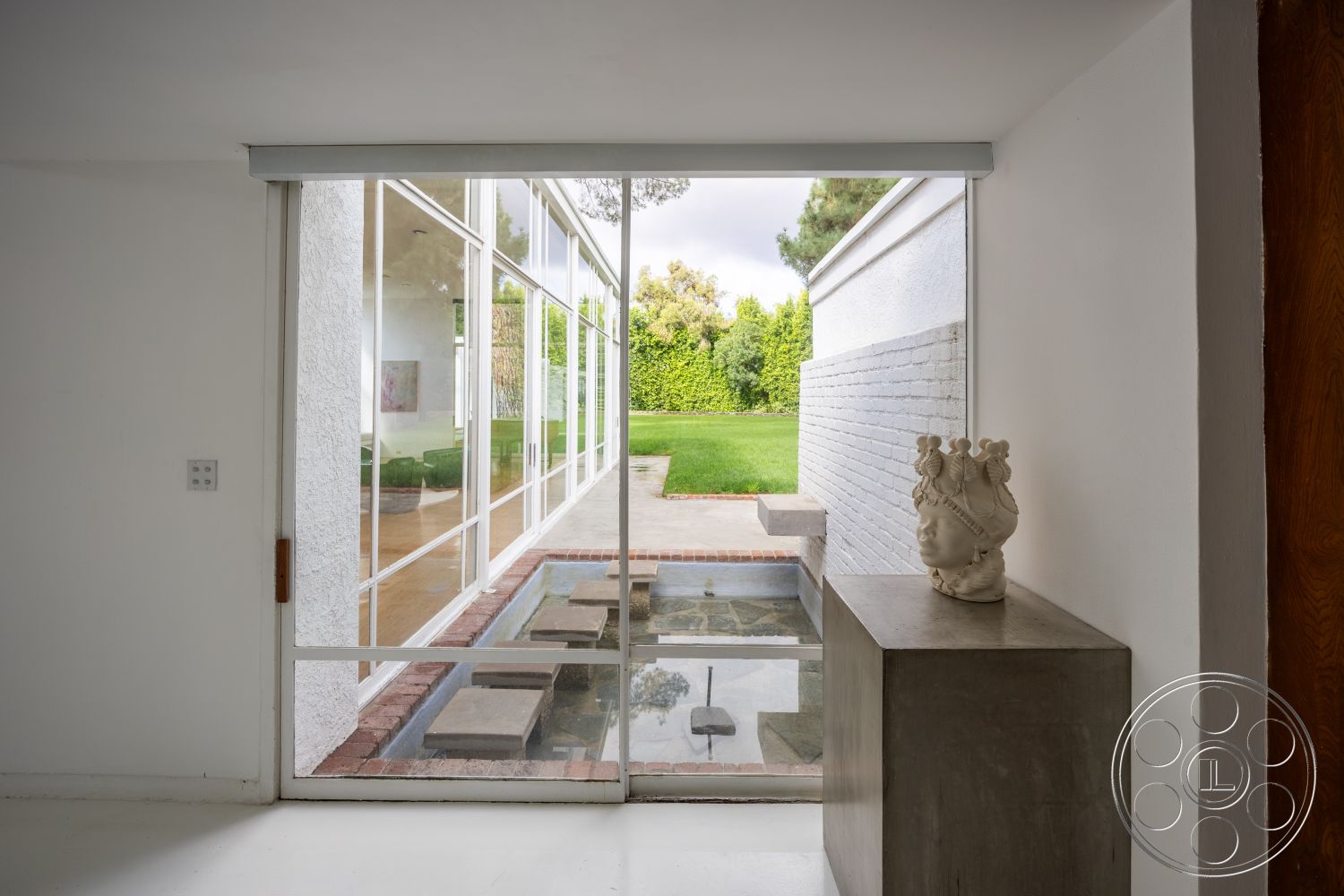 Sinatra House - landscaped garden view, vaulted ceiling, seamless indoor-outdoor flow, modernist design elements, light wood ceiling beams, open living space layout, concrete feature wall, interior glass walls, natural light from floor-to-ceiling windows, contemporary interior details, exterior white painted brick walls, rectangular pond with stone pavers, modern sculpture as interior decor, concrete countertop pedestal, minimalist architecture design, green lawn setting