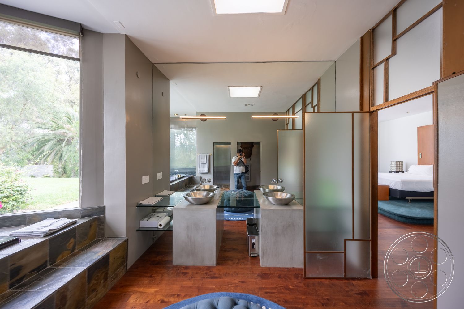 Sinatra House - mid-century modern architectural style, interior bathroom with polished concrete countertops, wood beam ceiling accent in bedroom area, floating glass shower doors in modern bathroom, dark walnut hardwood flooring in bedroom, stainless steel sink bowls in dual vanity, slate stone seating area by large window, modern minimalist design with frosted glass partitions, large picture windows with garden view, open-concept layout with seamless transition from bathroom to bedroom