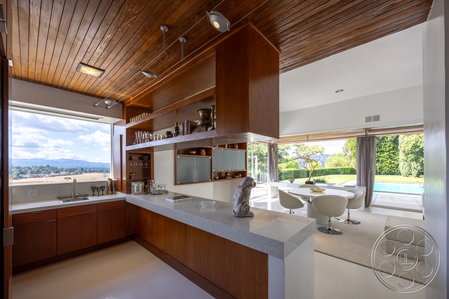 Sinatra House - open-concept living space, floor-to-ceiling windows in dining area, exterior kitchen with panoramic mountain views, wood ceiling beams in kitchen, concrete countertop with glossy finish, modern wooden cabinetry, glass sliding doors for indoor-outdoor flow, minimalist dining area with circular white table, grazing lawn with landscaped greenery, tile flooring in kitchen with seamless transitions, natural light flooding the interior, curved modern dining chairs, outdoor swimming pool visible from interior, light gray wall paint in kitchen, smooth concrete floor in dining area, landscape view through large glass panes