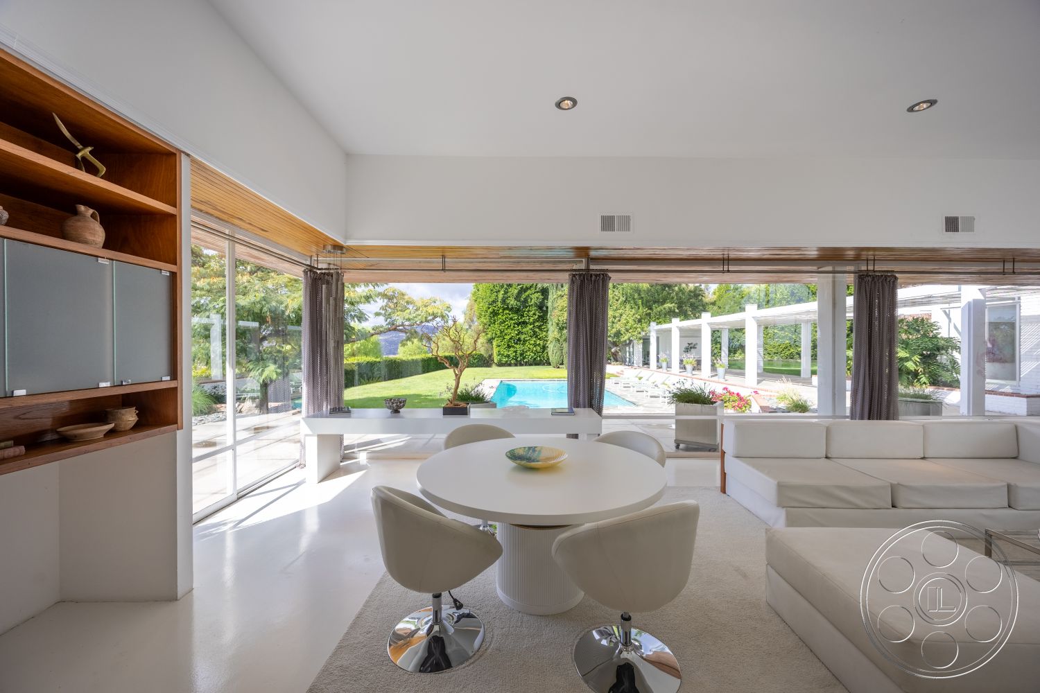 Sinatra House - landscaped garden, minimalist design, bright natural light, textured accent wall, glass sliding doors, floor-to-ceiling windows, open-concept layout, urban residential setting, curved pool shape, modern round dining table, wood beam ceiling, white painted walls, concrete patio floor, upholstered seating area, exterior pool view, integrated outdoor living