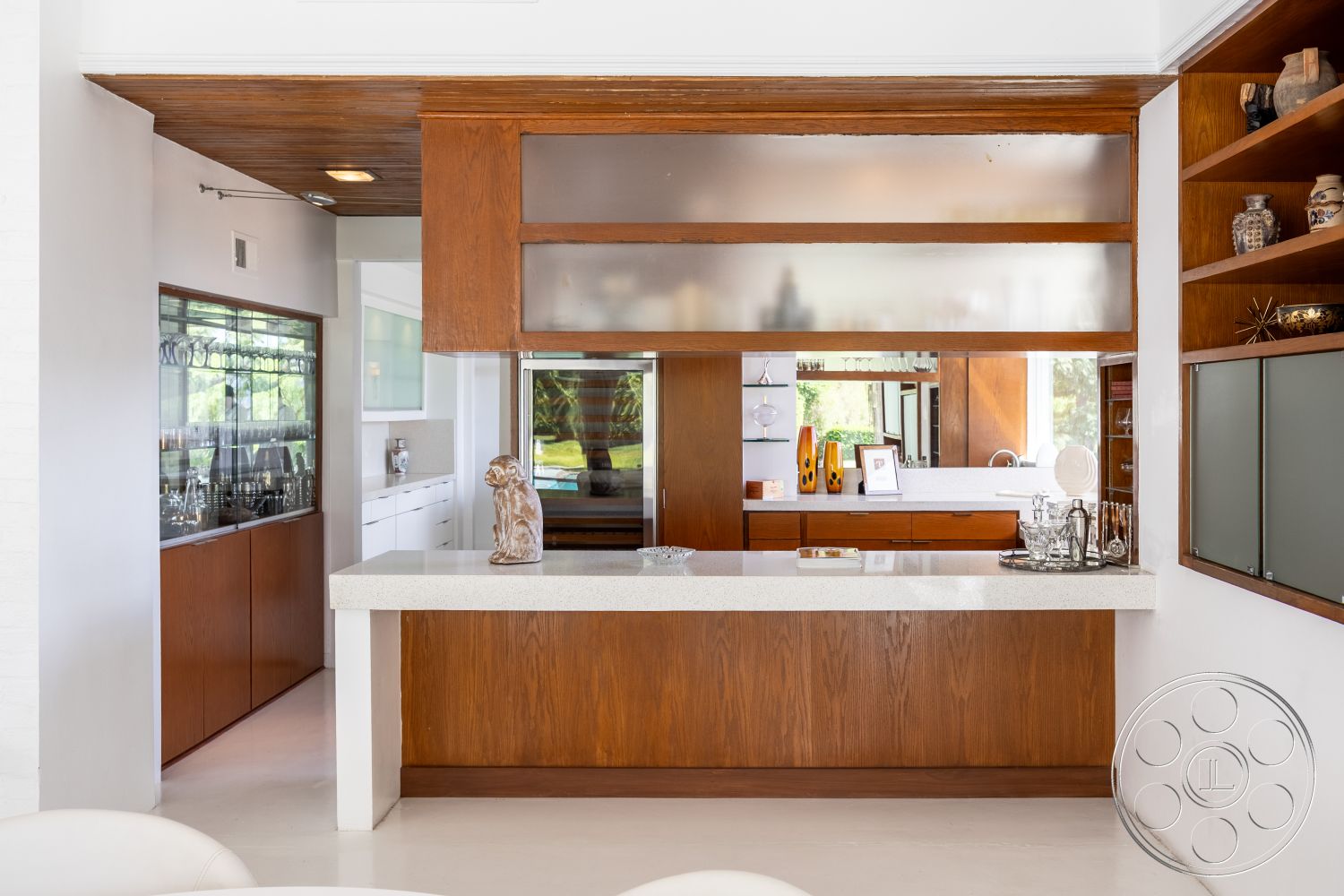 Sinatra House - natural light from large windows, textured plaster walls, mid-century modern architectural style, wood beam ceiling details, interior kitchen with quartz countertop, glass-front kitchen cabinets, walnut cabinetry in modern kitchen, open floor plan with visible kitchen layout, neutral tile flooring in kitchen, built-in shelving displays, frosted glass kitchen partition, minimalistic bar area, contemporary decor accents, seamless transition between living spaces