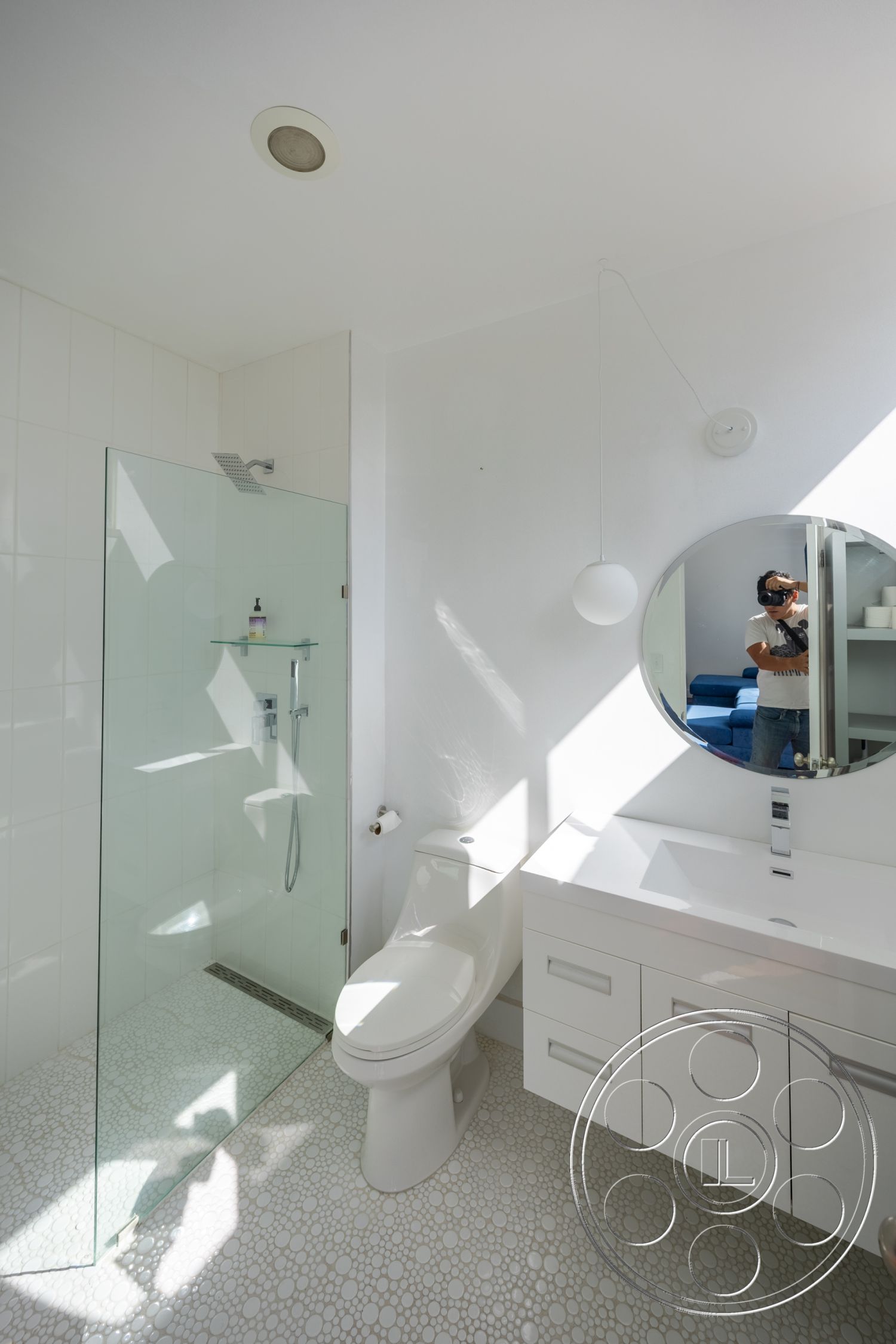 Sinatra House - neutral color palette, glass shower enclosure, natural light from window, minimalist bathroom design, interior bathroom, white tile shower walls, porcelain pebble floor tiles, floating vanity with white cabinetry, round mirror bathroom fixture, chrome bathroom faucet, backlit vanity lighting, modern plumbing fixtures